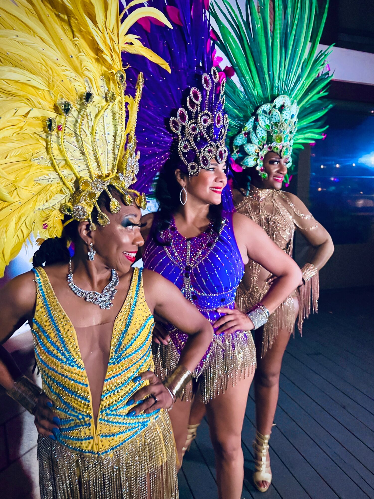 Three women in colorful carnival costumes with large feathered headdresses, posing together indoors.