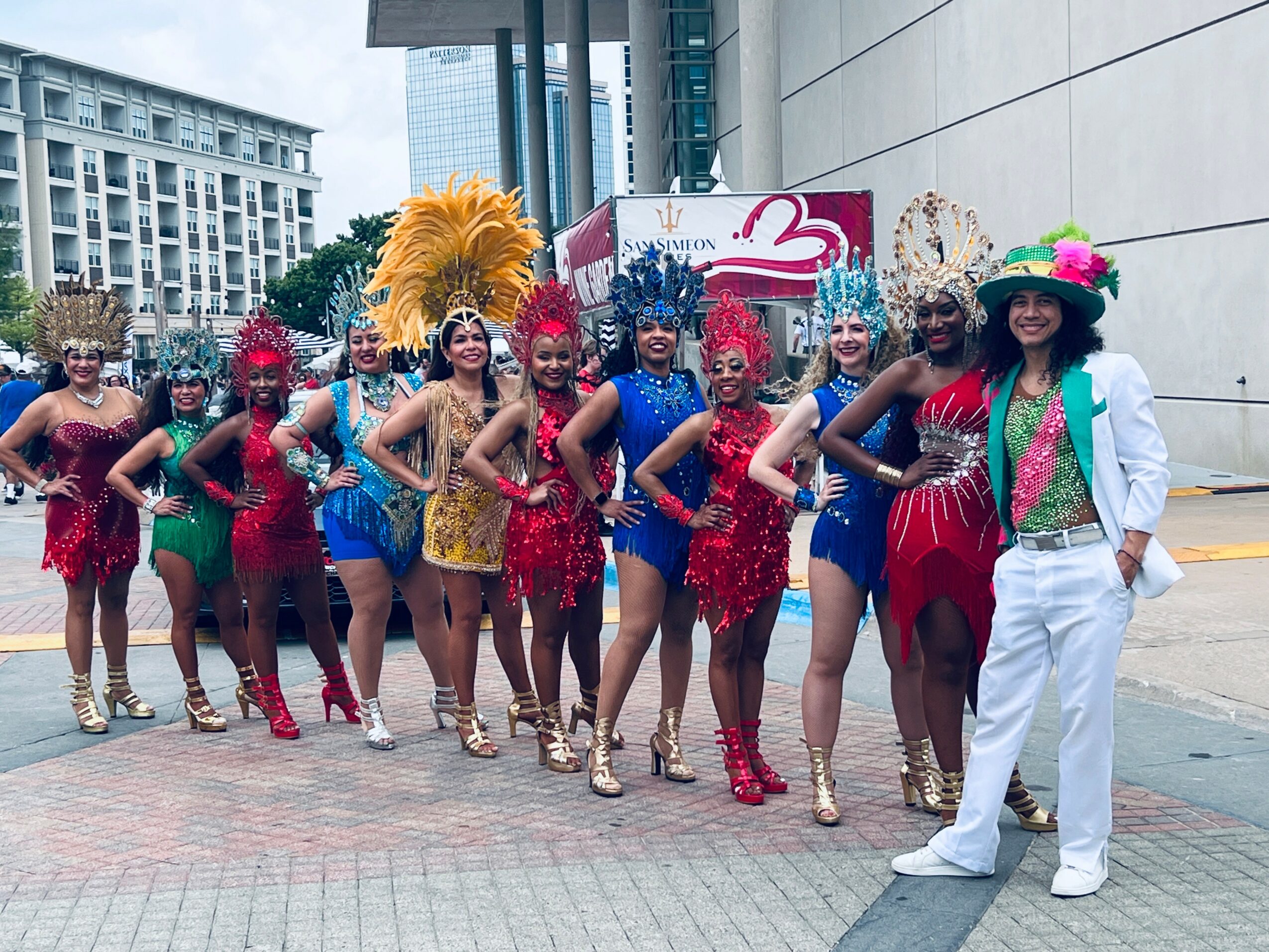 Group of women in colorful costumes and a woman in white suit and hat standing outdoors in an urban area.