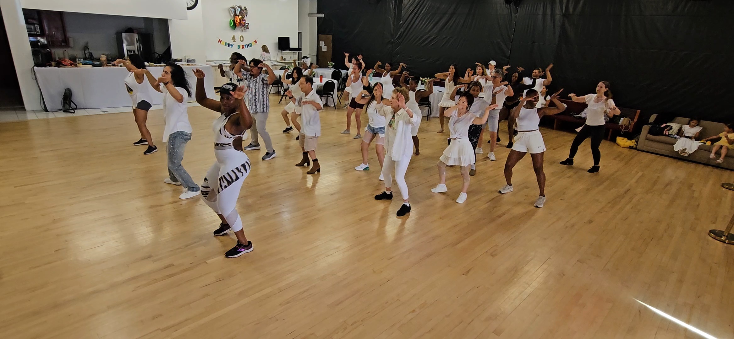 Group of children dancing in a spacious room with wooden floor and black wall, some wearing white clothes, some striped.