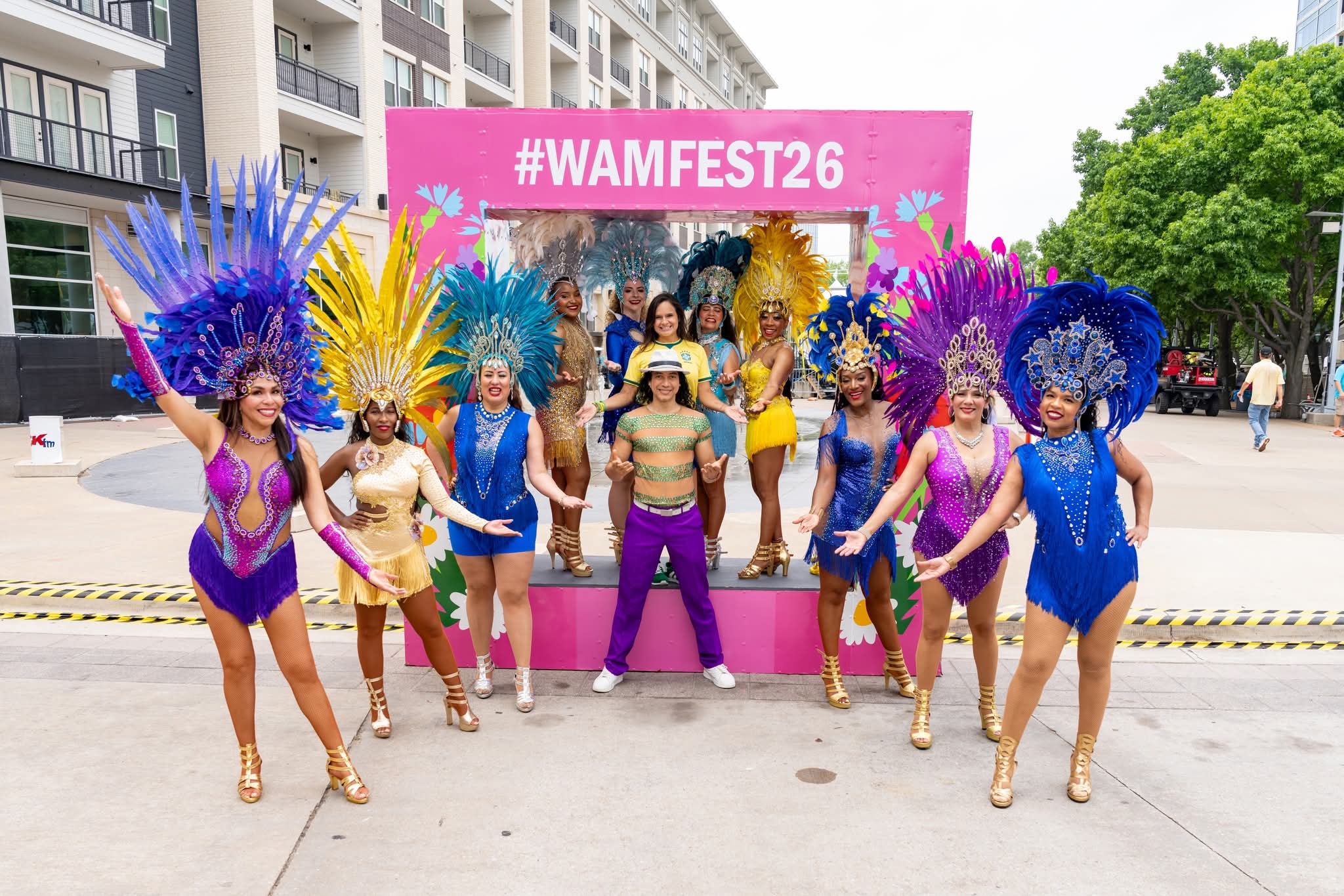 Group of performers in colorful costumes and feathered headdresses posing outdoors in front of a pink banner with #WAMFEST26, on a city street.
