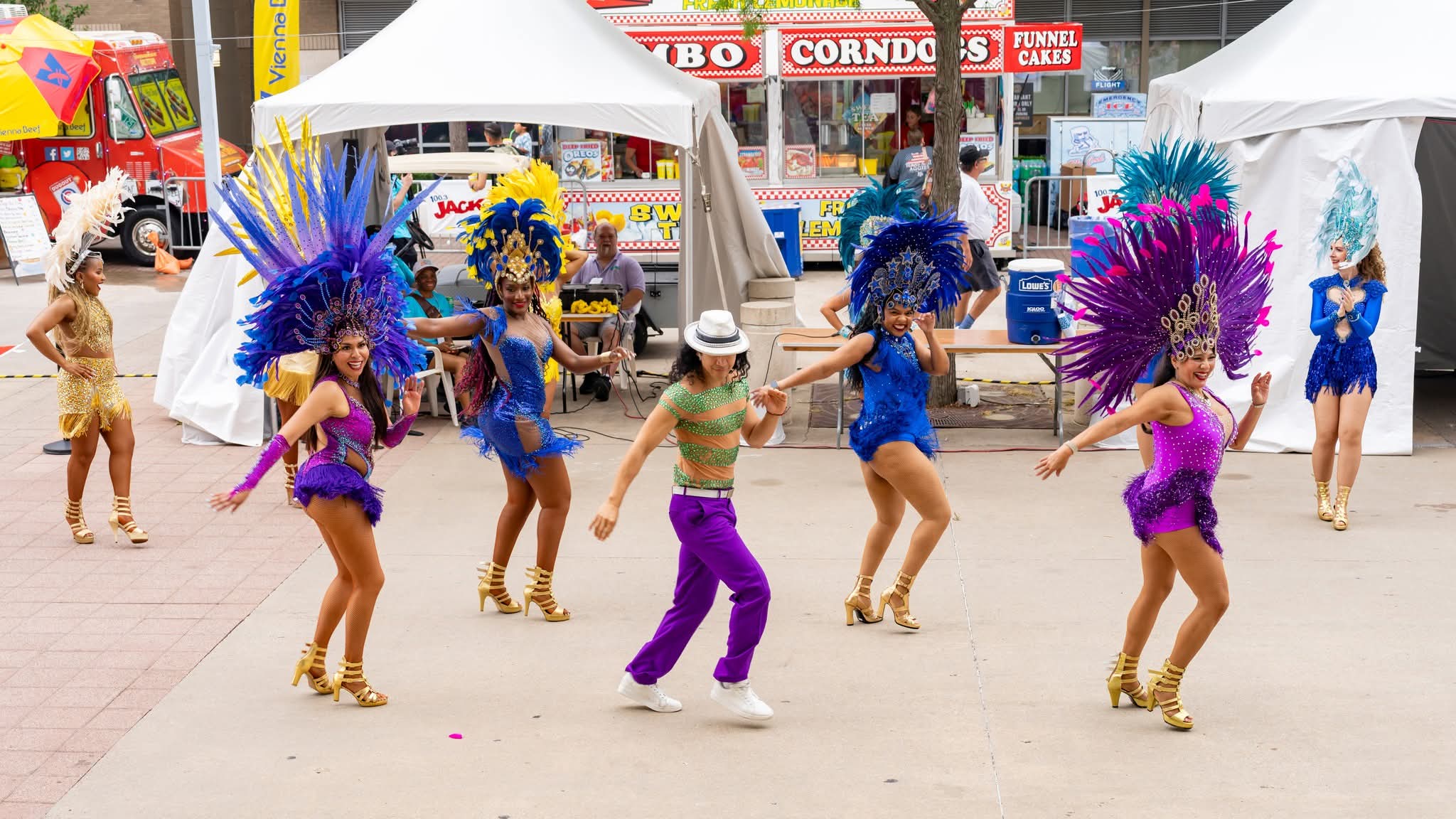 Group of dancers in colorful costumes performing outdoors with tents and market stalls in background.