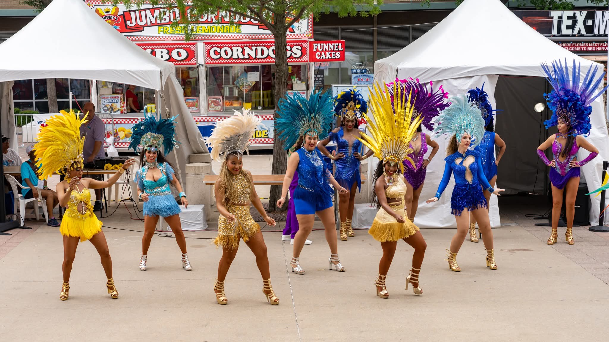 Group of dancers in colorful costumes with large feathered headpieces performing outdoors, tents and signs in background.