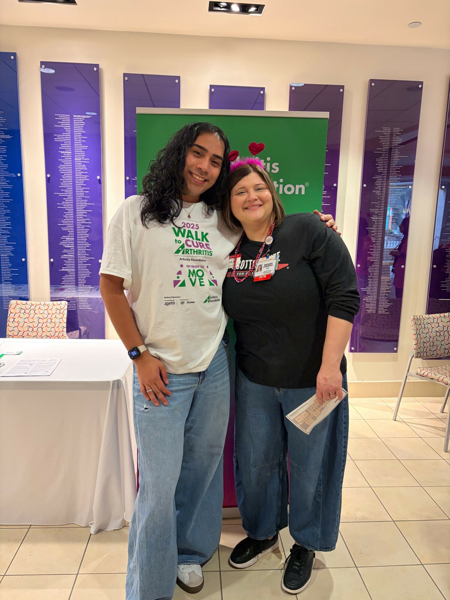 Two women standing indoors in front of a purple and green display, smiling at the camera, one wearing a white shirt and the other a black shirt.