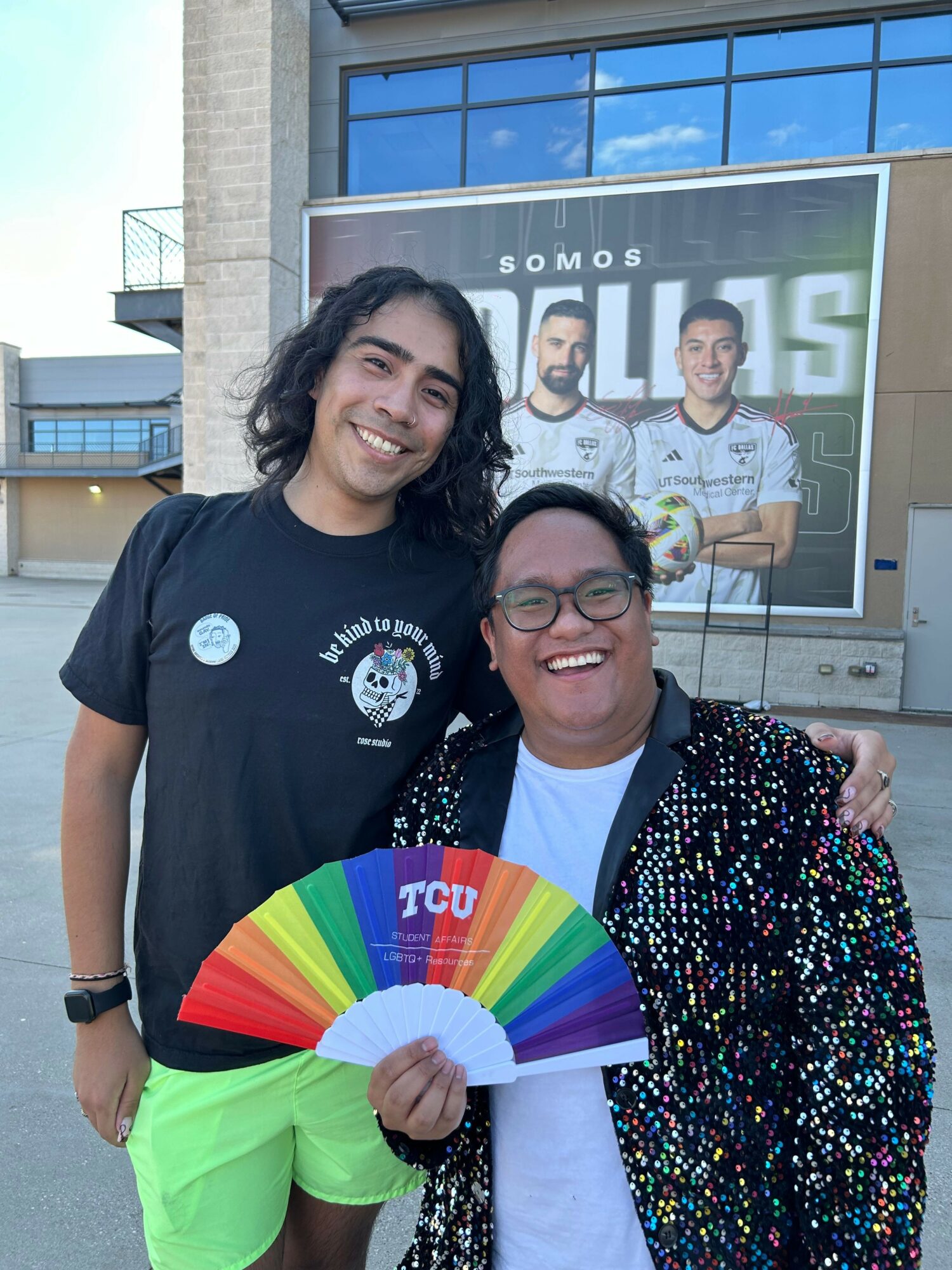 Two women smiling outside a building, one holding a rainbow fan, with a poster in the background.