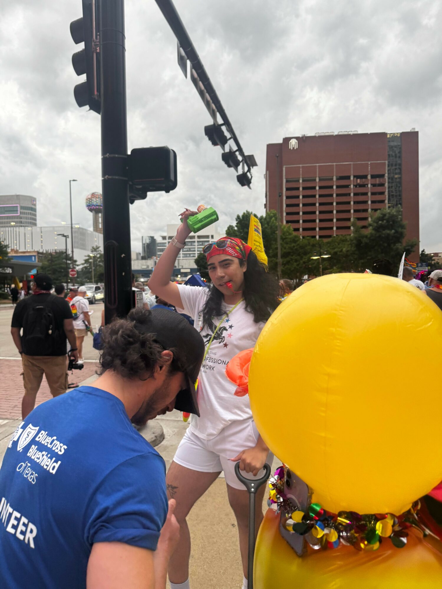 People gathered outdoors, some holding balloons, with buildings and cloudy sky in the background.