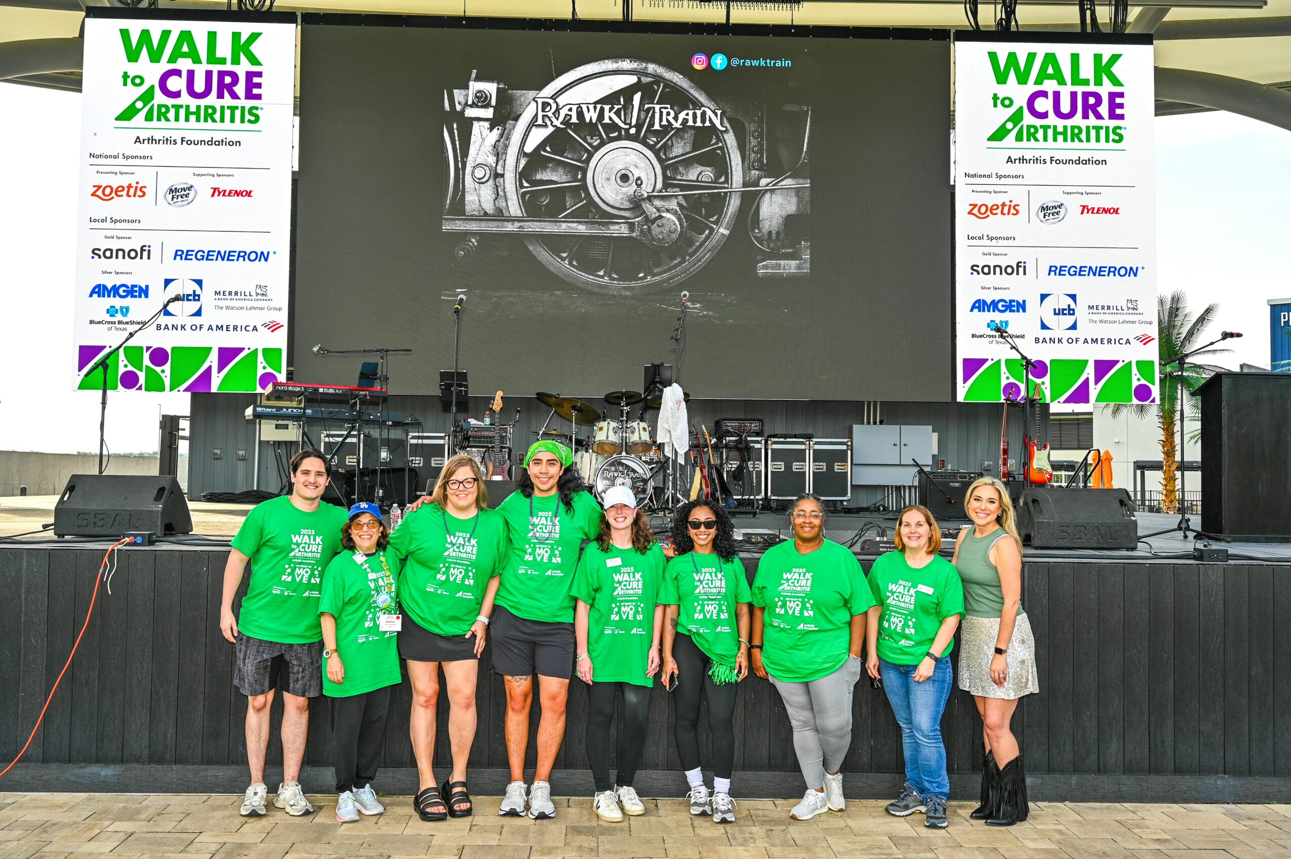 Group of seven people standing in front of a stage with a large screen displaying 'Walk to Cure Arthritis' logo, all wearing green shirts.