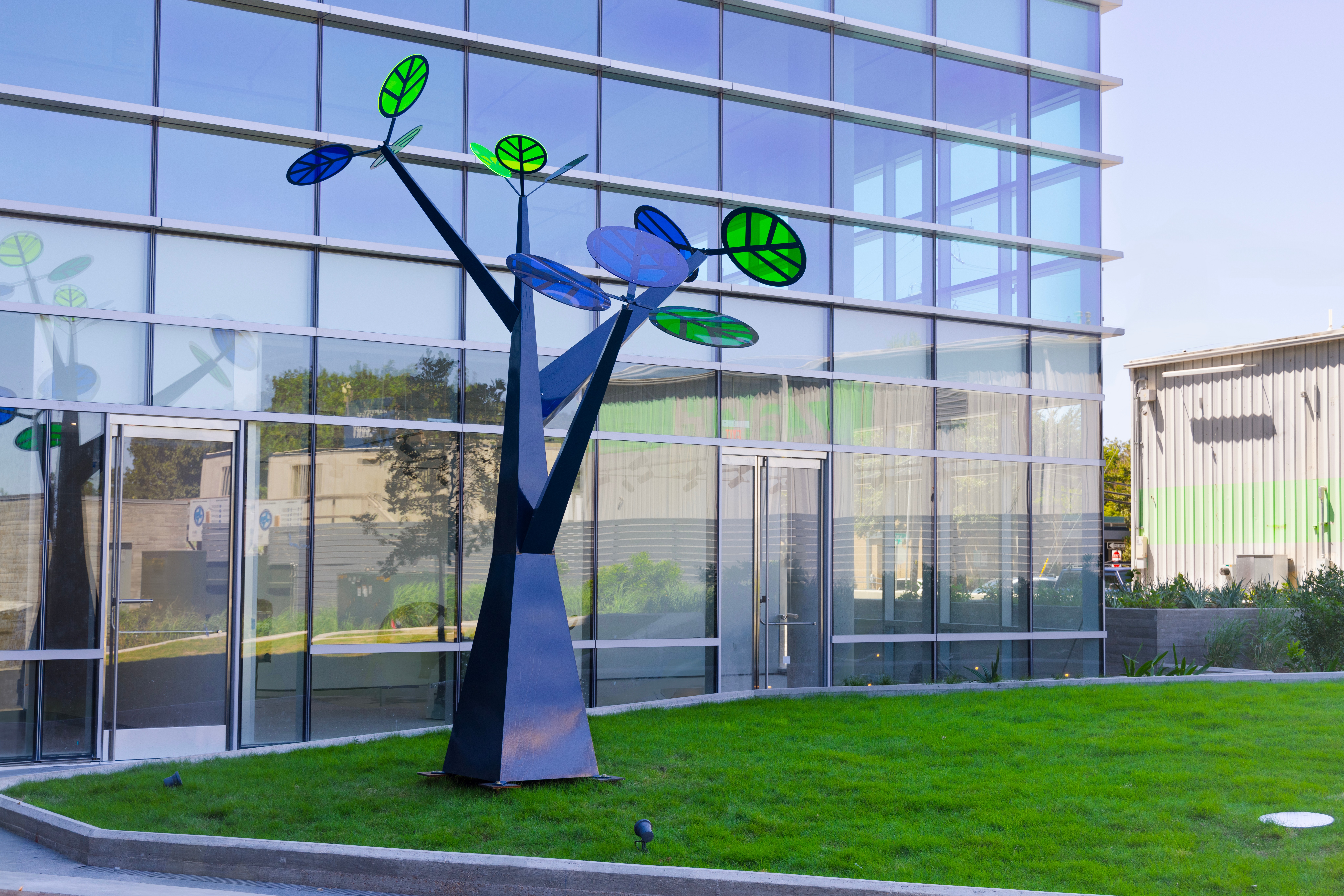 Modern sculpture of a person with outstretched arms holding green leaves, outside a glass building, on a grassy area.