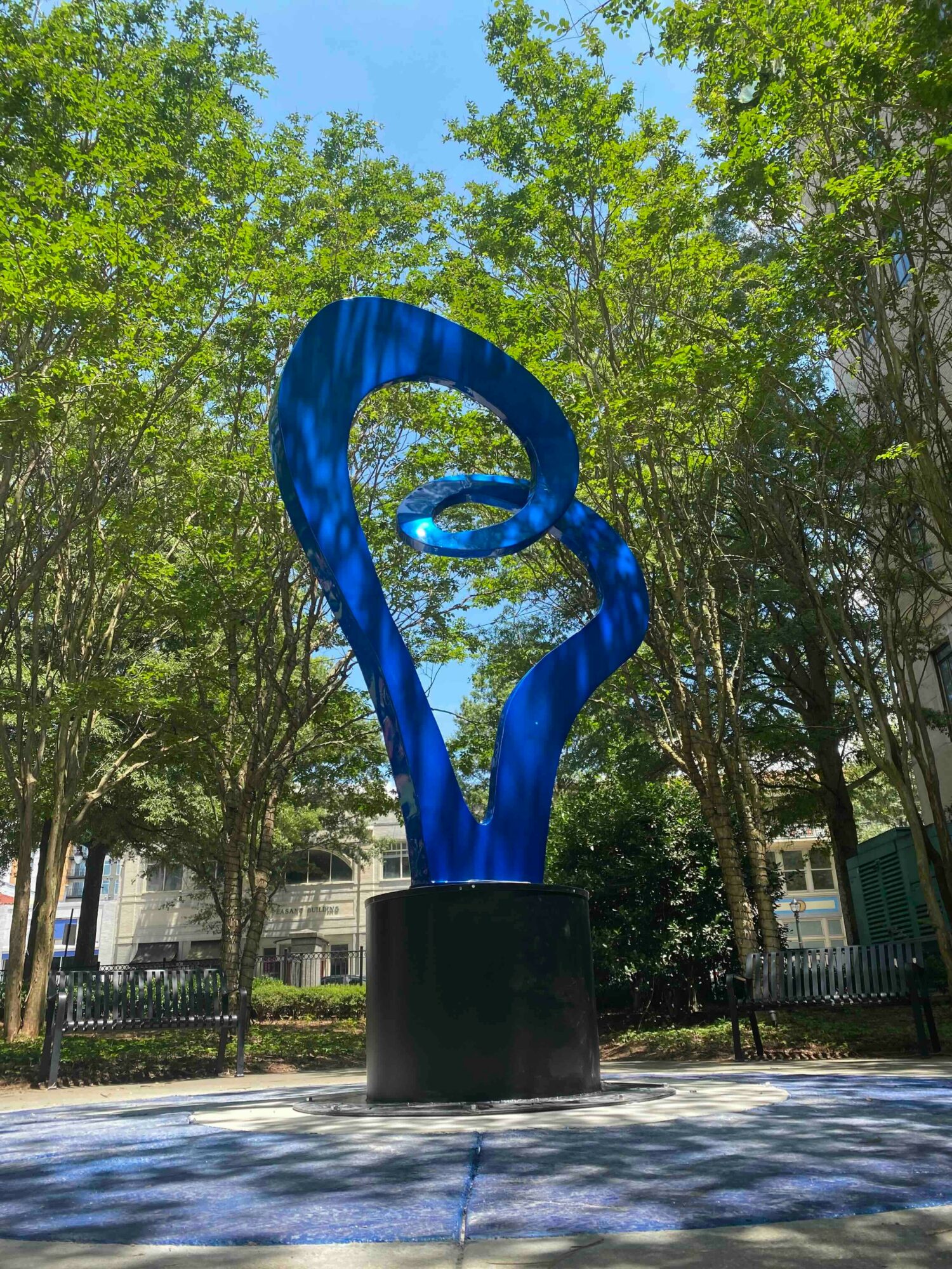 Blue abstract sculpture in a park with trees and benches, sunlight filtering through leaves.