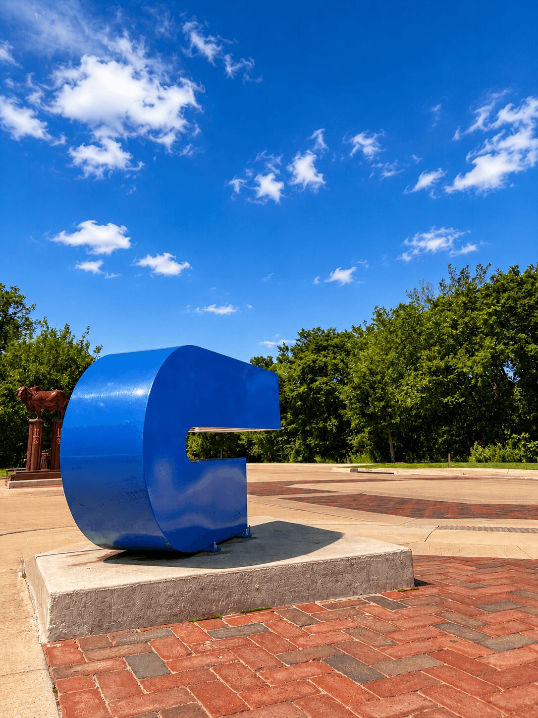 Large blue letter G sculpture on a concrete platform outdoors, with trees and a blue sky with clouds in the background.