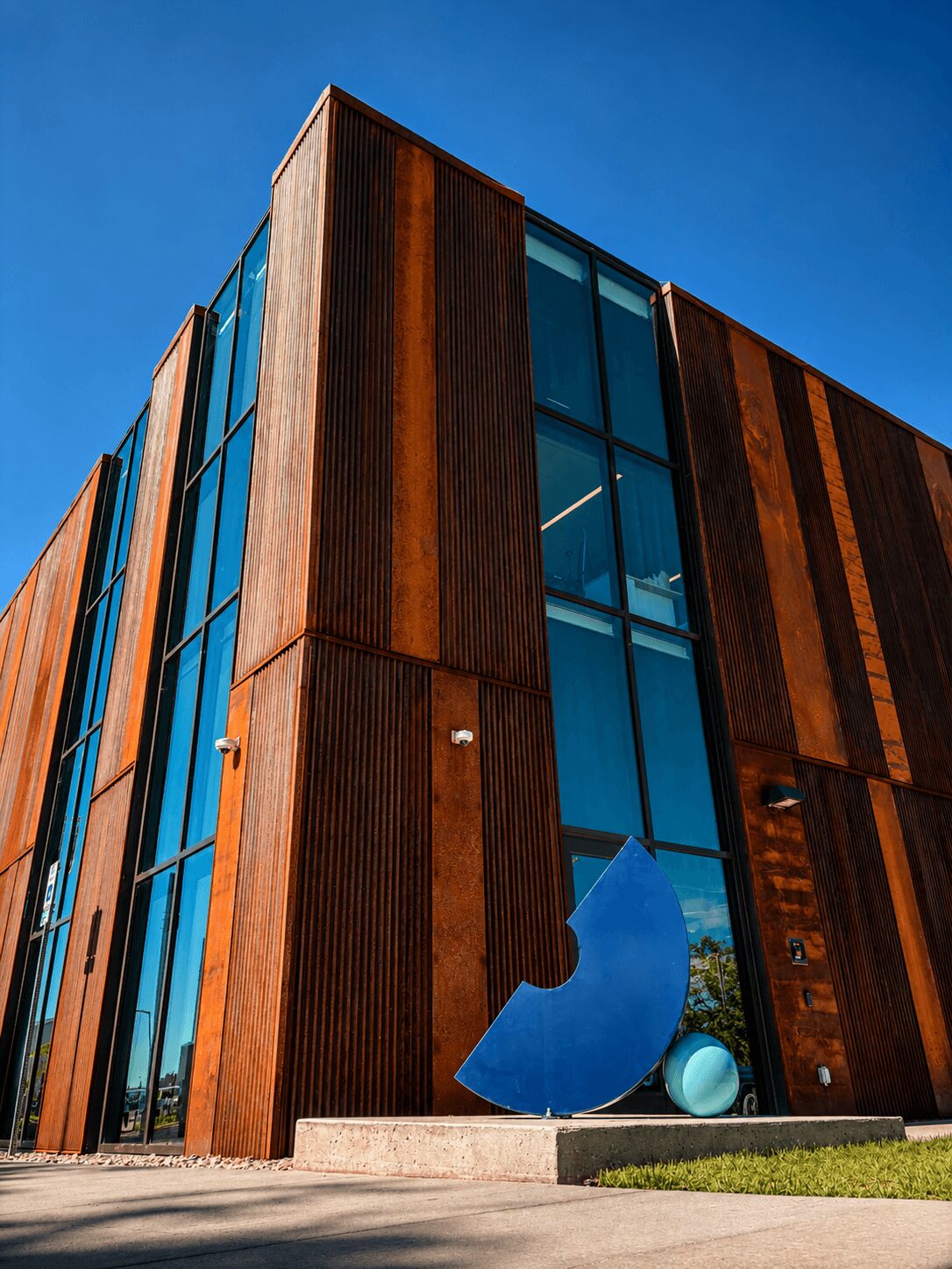 Modern building with large glass windows and rust-colored metal panels, featuring a blue sculpture outside, against a clear blue sky.