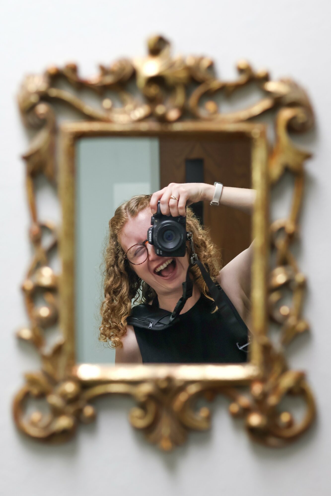 Person with curly hair and glasses taking a photo in a mirror, smiling, with an ornate gold frame, background blurred.