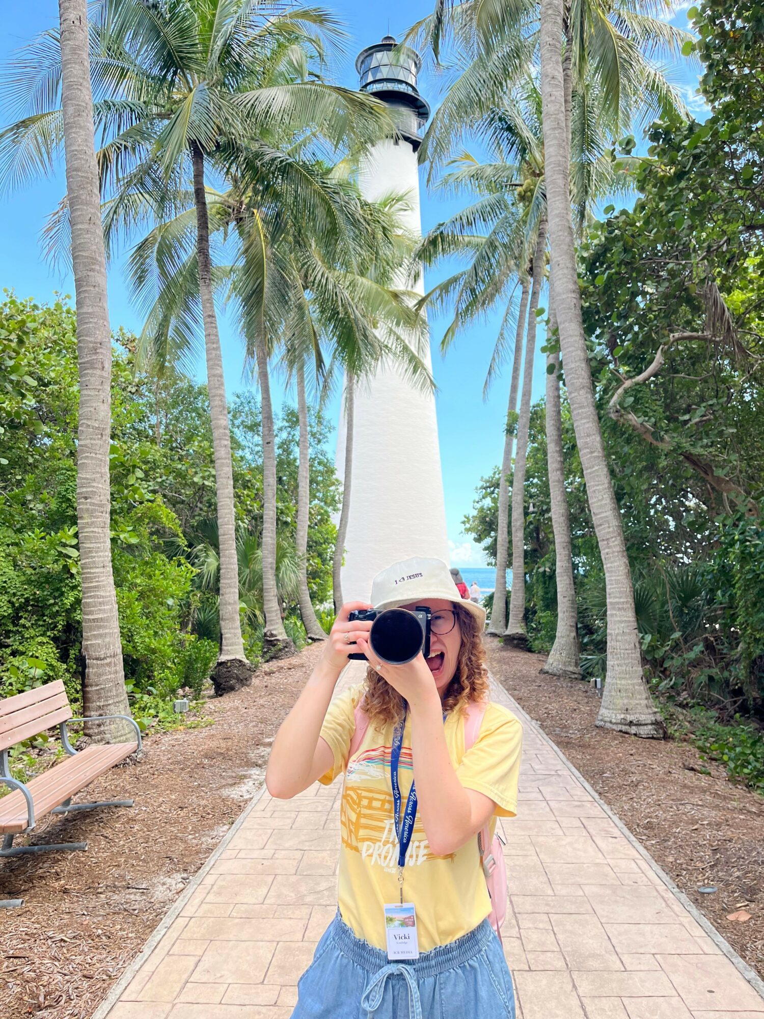 Person taking a photo with a camera on a sidewalk lined with palm trees, blue sky in background.