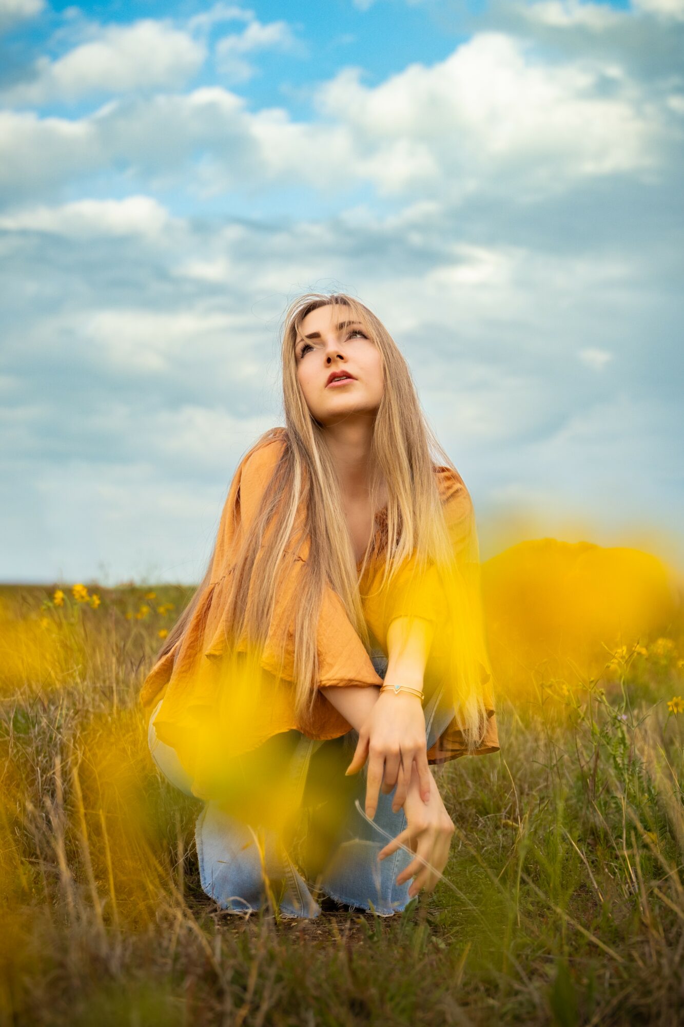 Young woman with long hair kneeling in a field of yellow flowers under a cloudy sky, looking upward.