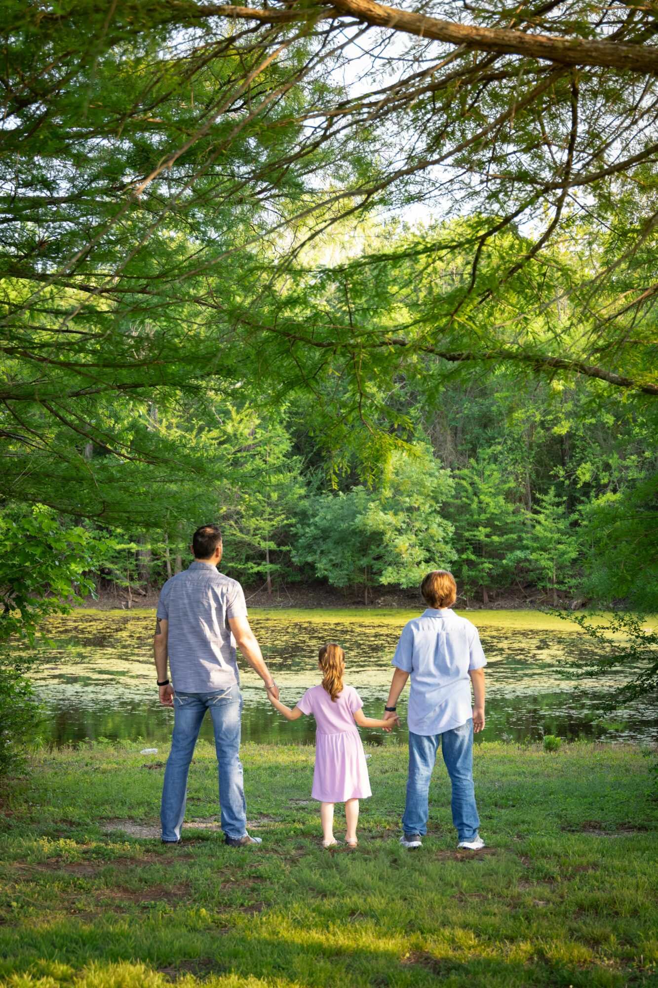 Three people, two adults and a child, holding hands and walking in a park with trees and greenery.