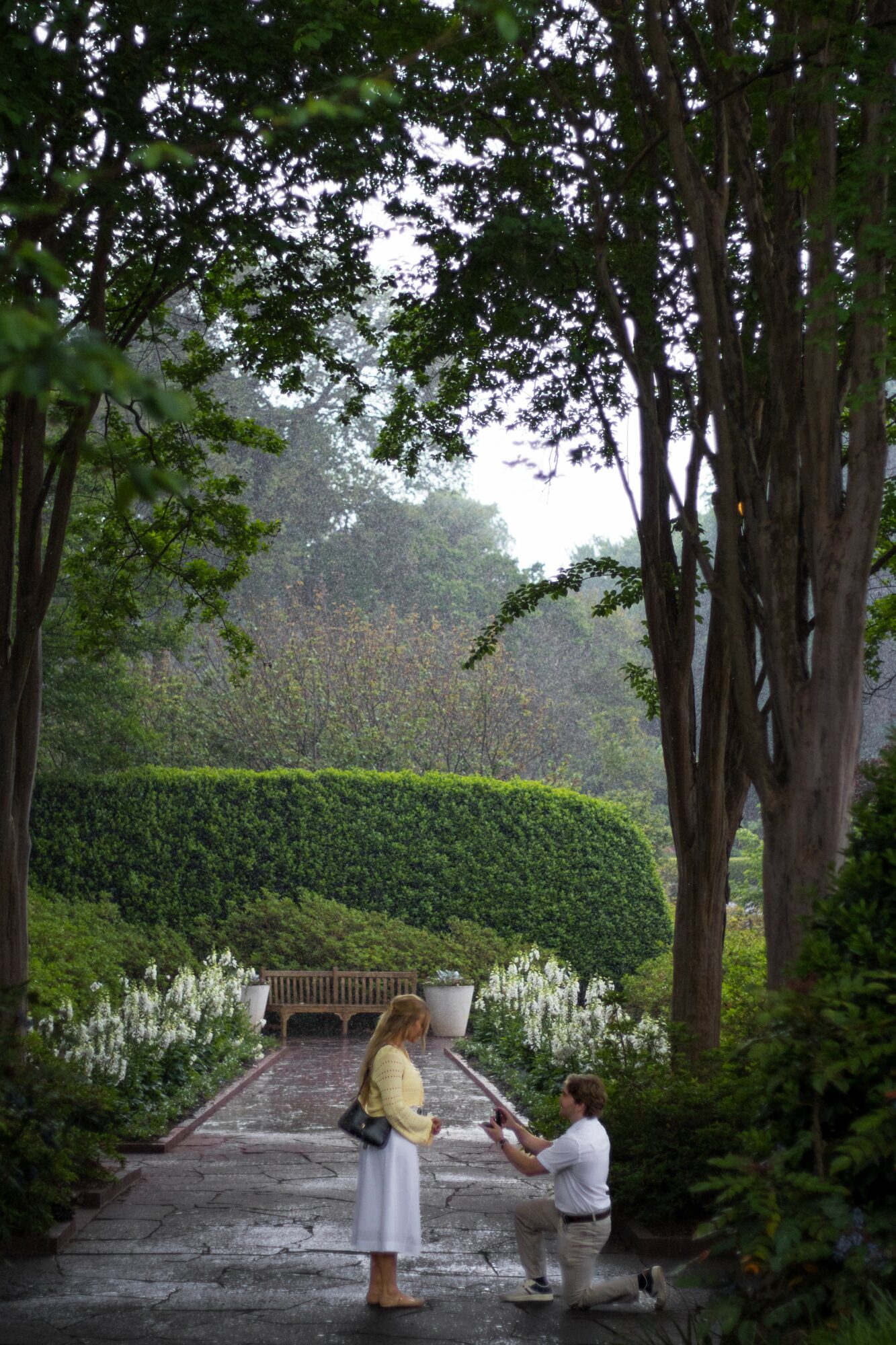 Person kneeling on a pathway holding hands with a child in a garden with trees and bushes.