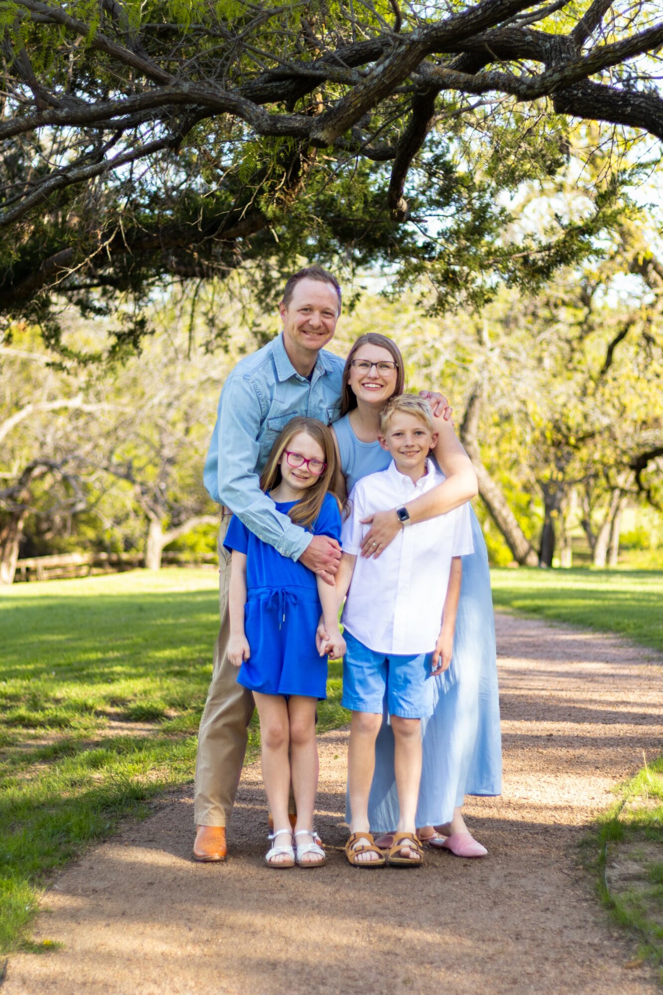 Family of four standing outdoors under a tree, smiling, with a park background, sunlight, and green grass.
