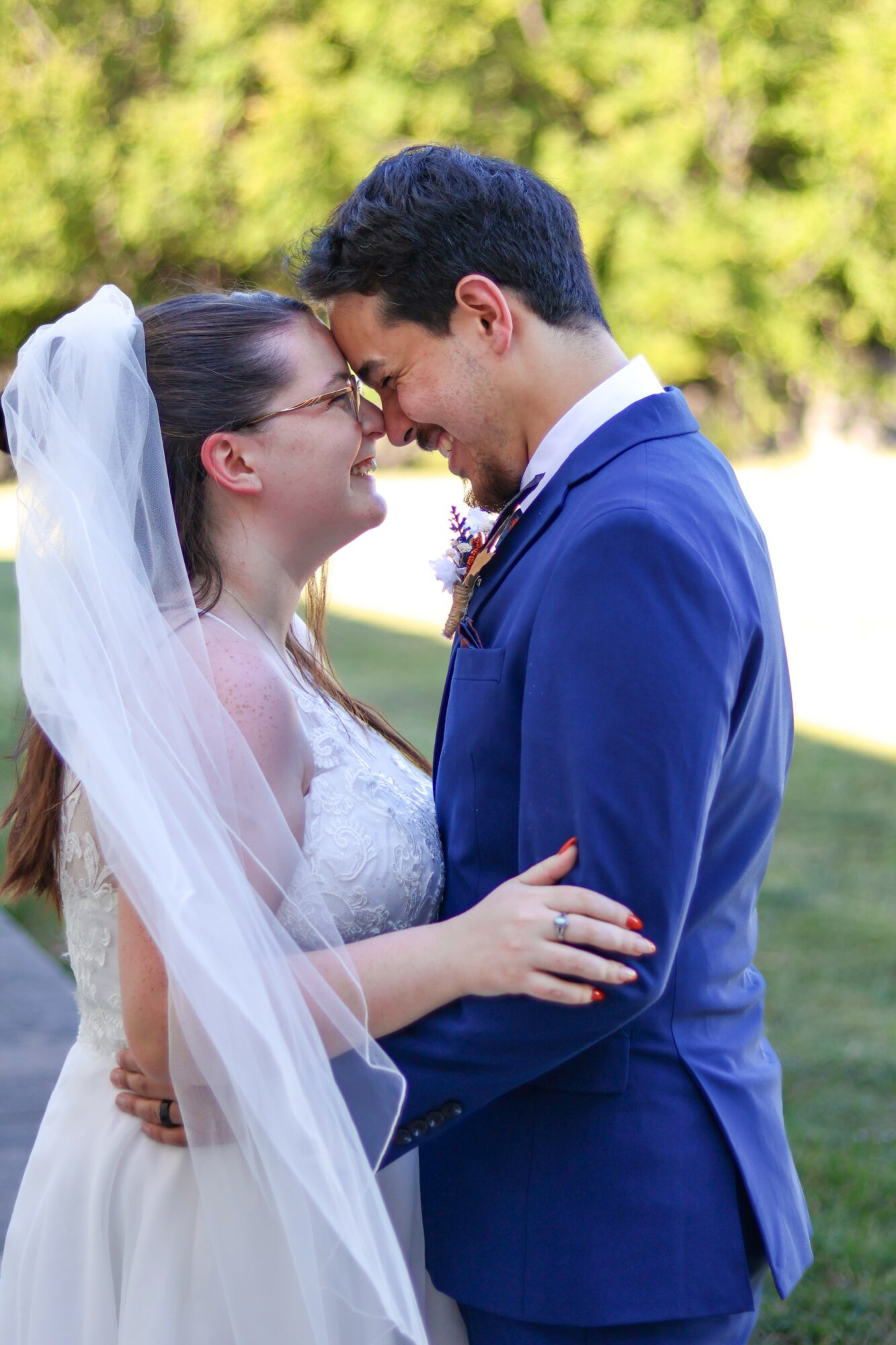 Bride and groom face each other outdoors, smiling, with trees in the background.