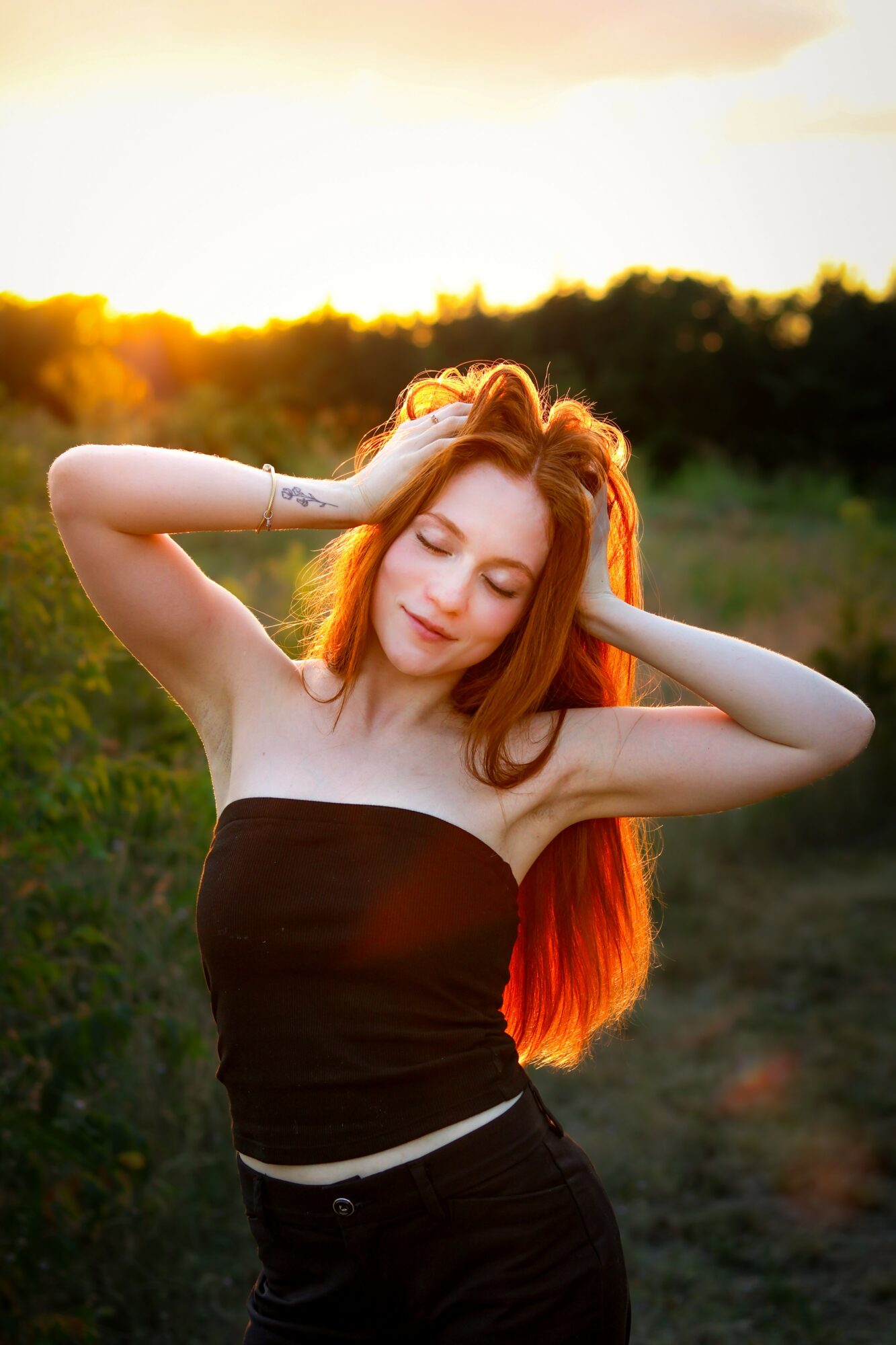 Young woman with long red hair, eyes closed, outdoors during sunset, wearing a black strapless top, hands in hair.