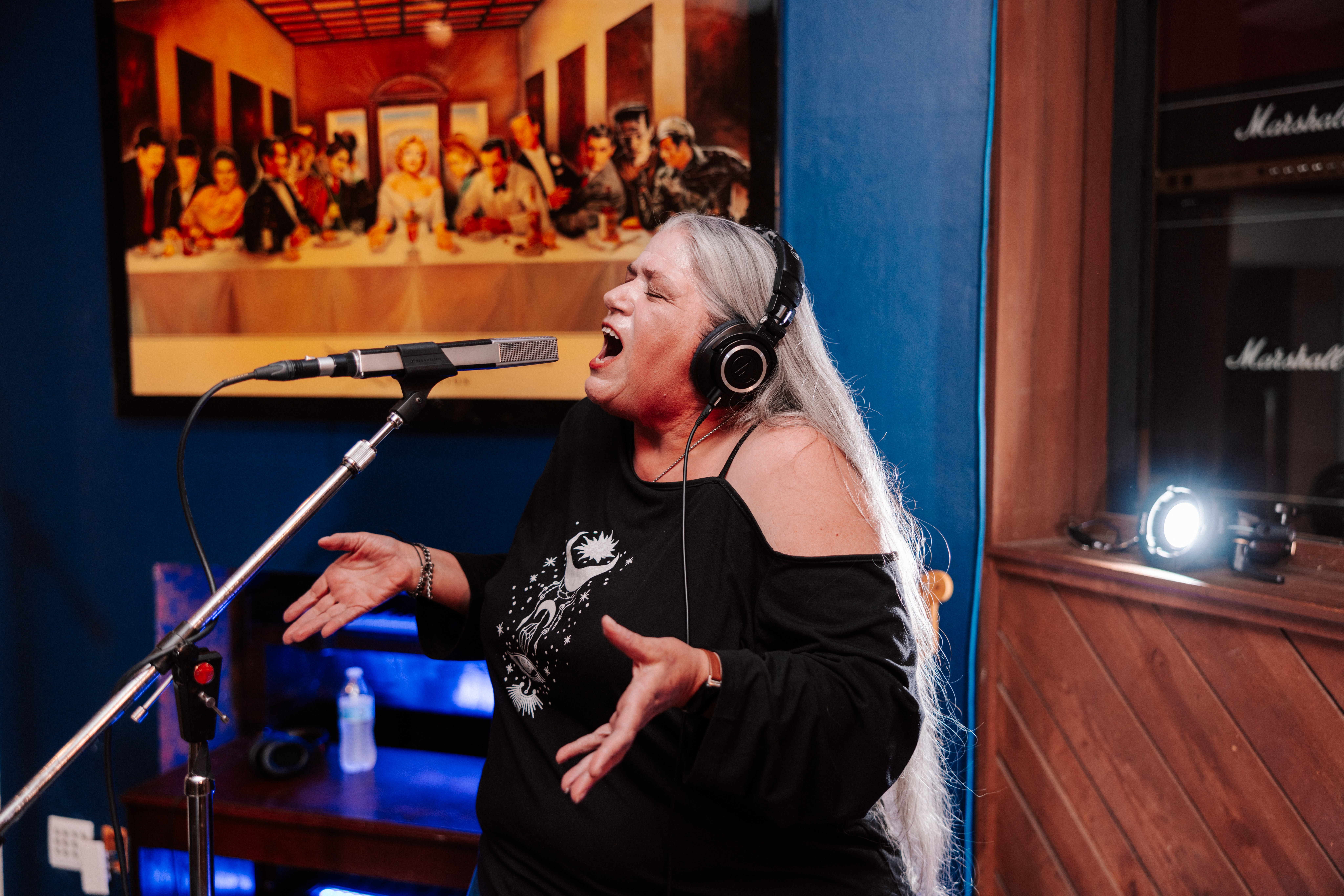 Woman singing into microphone with headphones, gesturing with hands, in recording studio, colorful painting behind her.