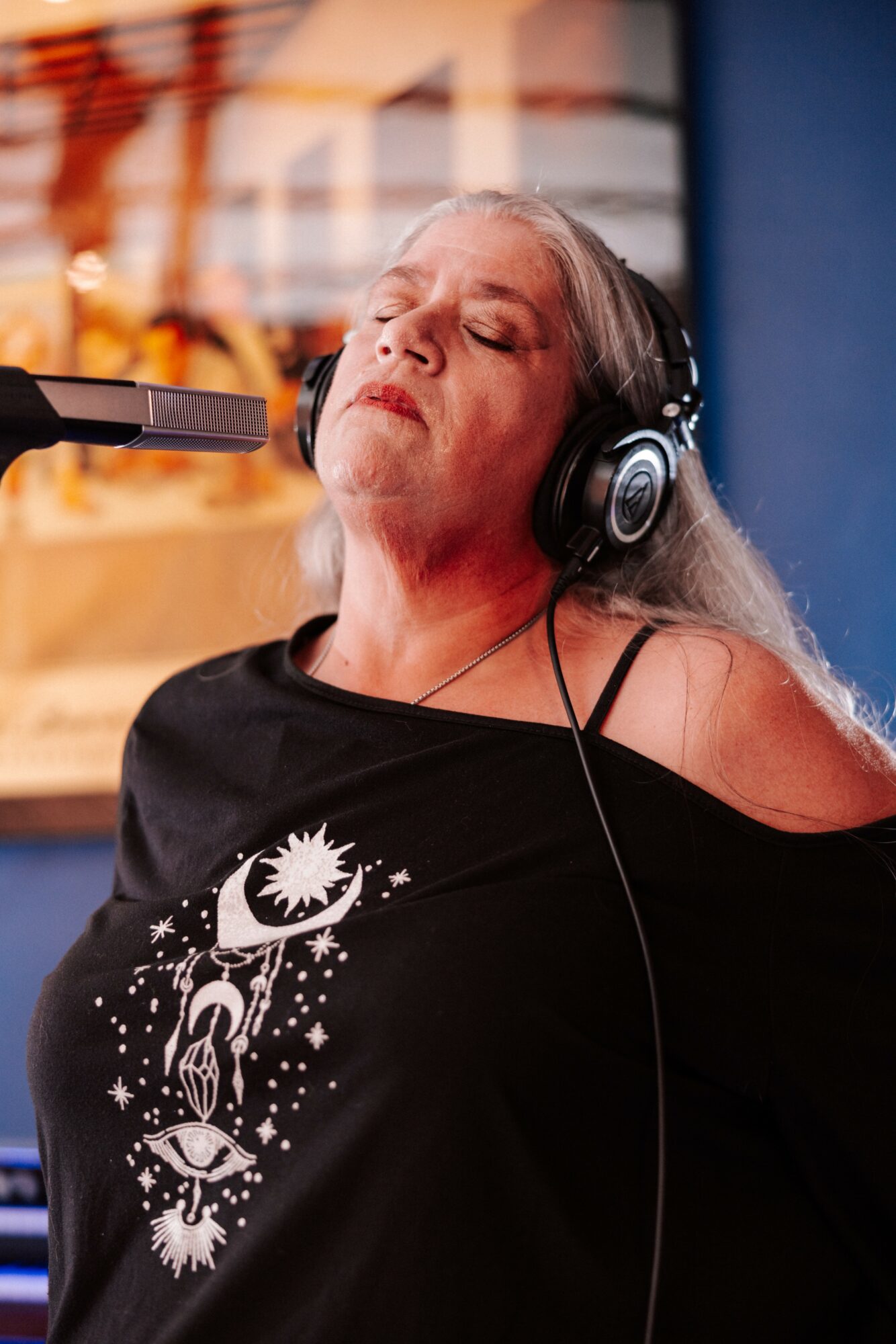 Elderly woman with long gray hair wearing headphones, eyes closed, listening to music, wearing a black shirt with celestial design.