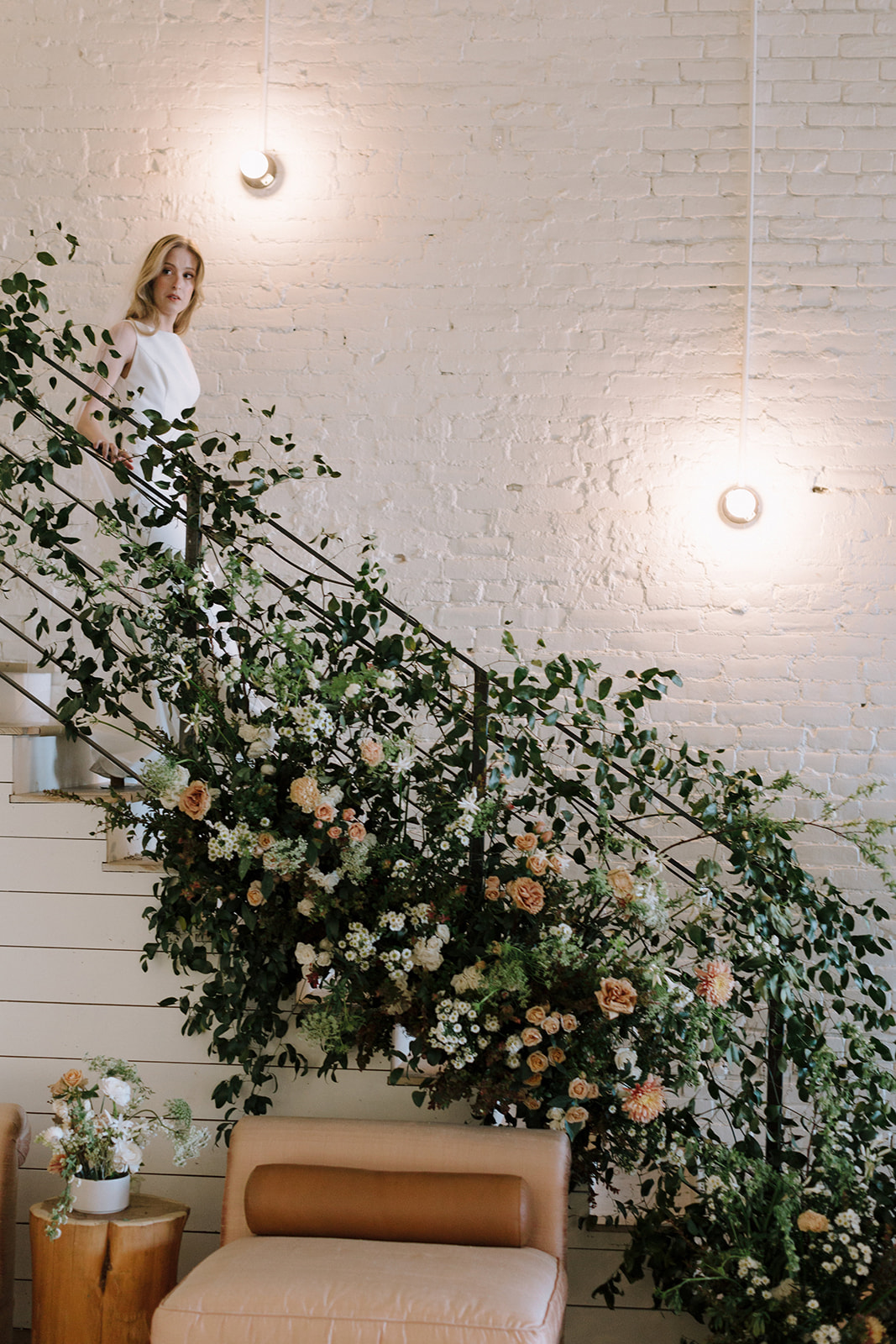 Woman standing on staircase next to floral arrangement and white brick wall with lights, indoor setting.