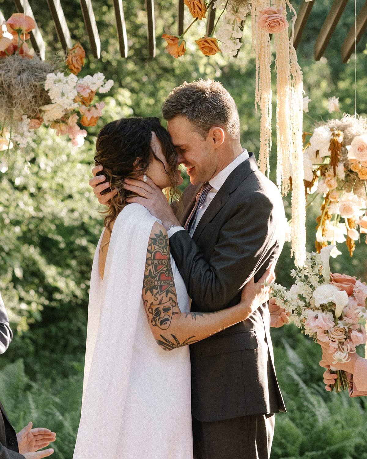 A couple embraces outdoors under floral decorations, smiling and looking at each other, with trees in the background.