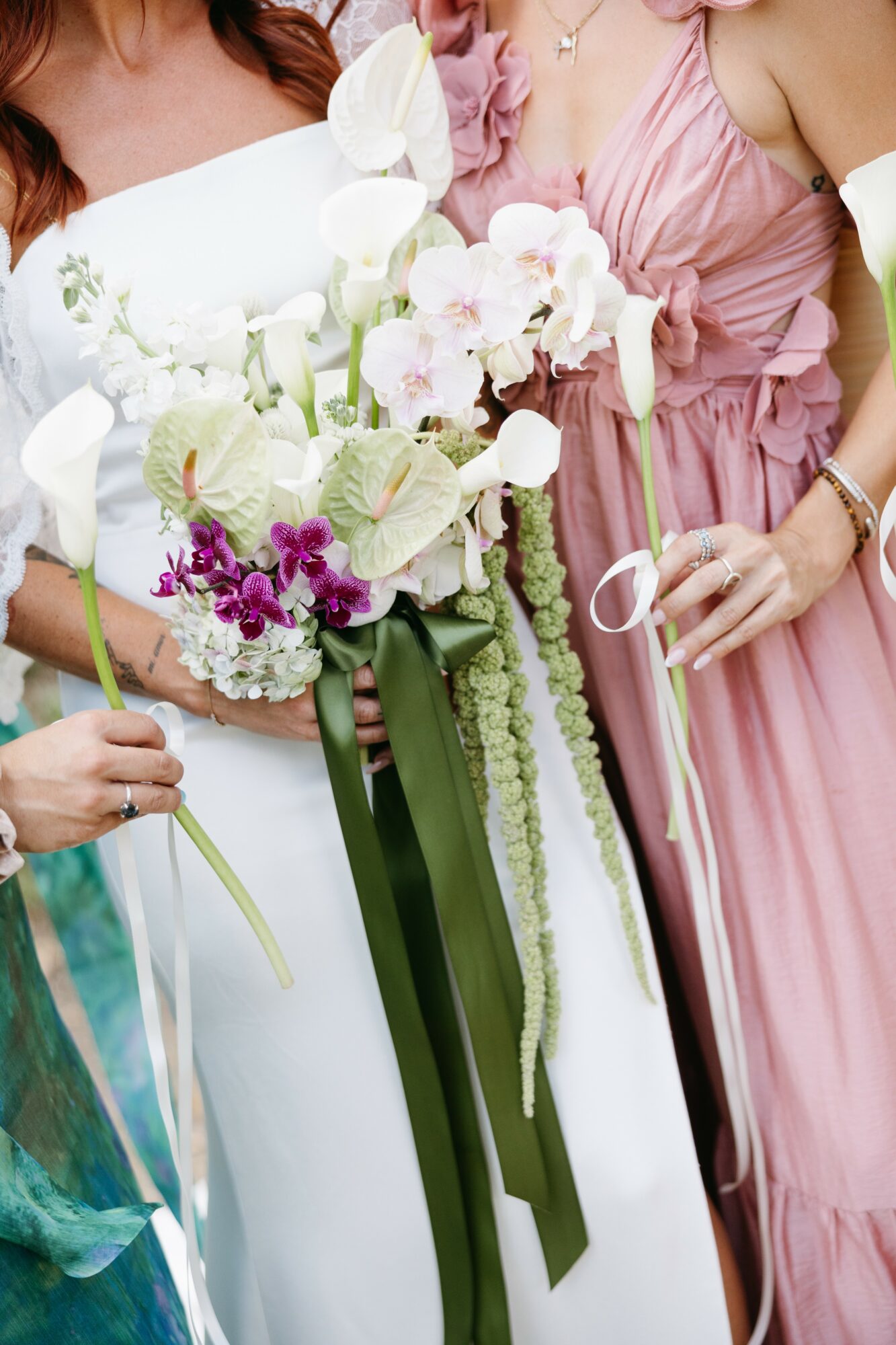 Two women in elegant dresses hold a large bouquet of white, pink, and purple flowers with green ribbons.