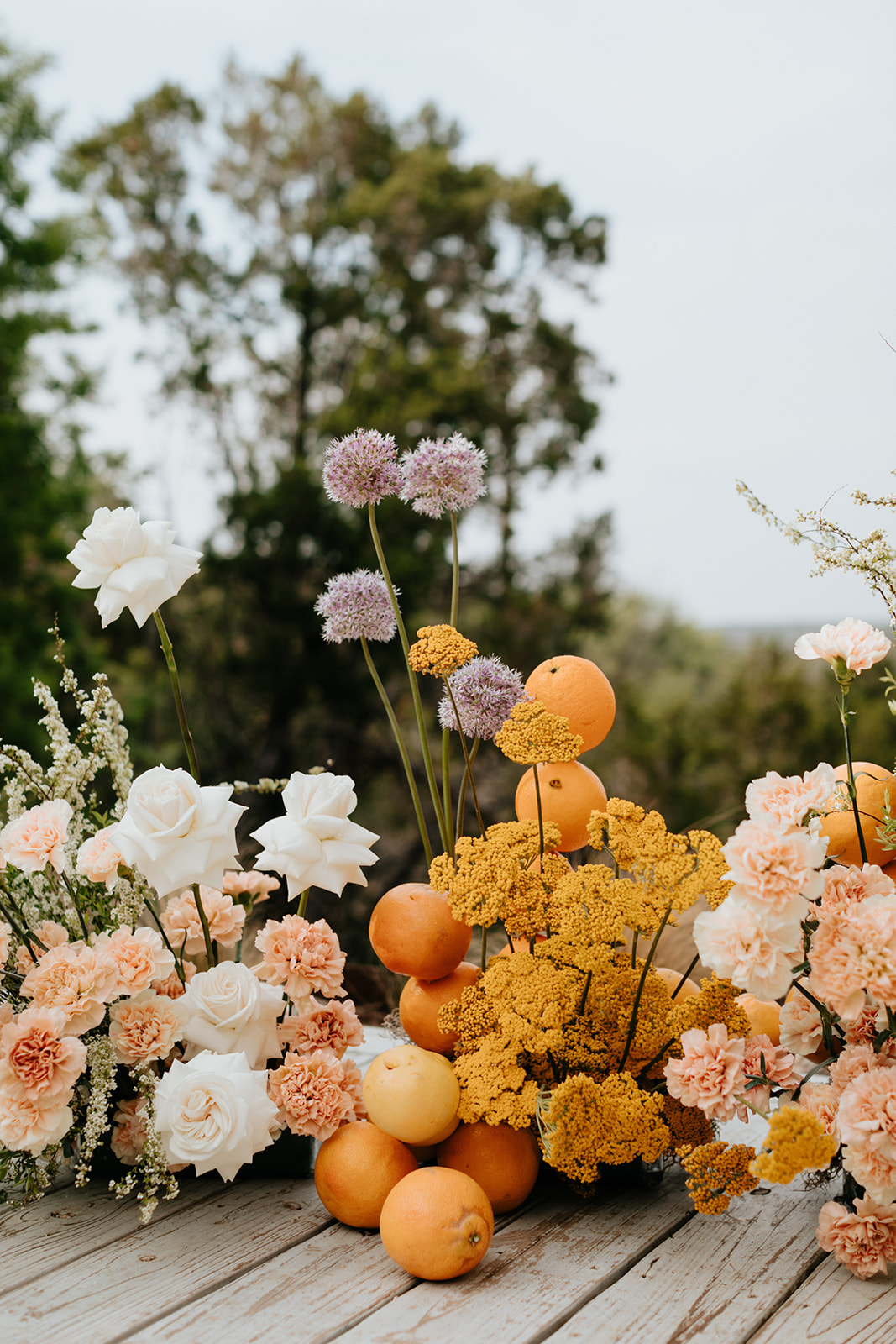 Arrangement of pink, white, and yellow flowers with oranges on a wooden surface outdoors.