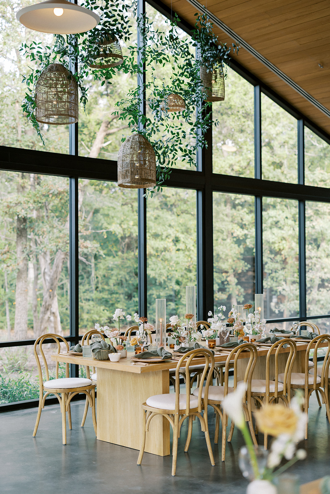 Dining area with large windows, wooden table, chairs, and hanging pendant lights, overlooking greenery outside.