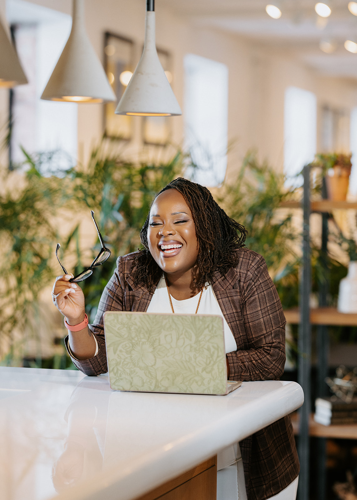 Smiling woman with glasses in hand, working on a laptop in a bright, plant-filled space.