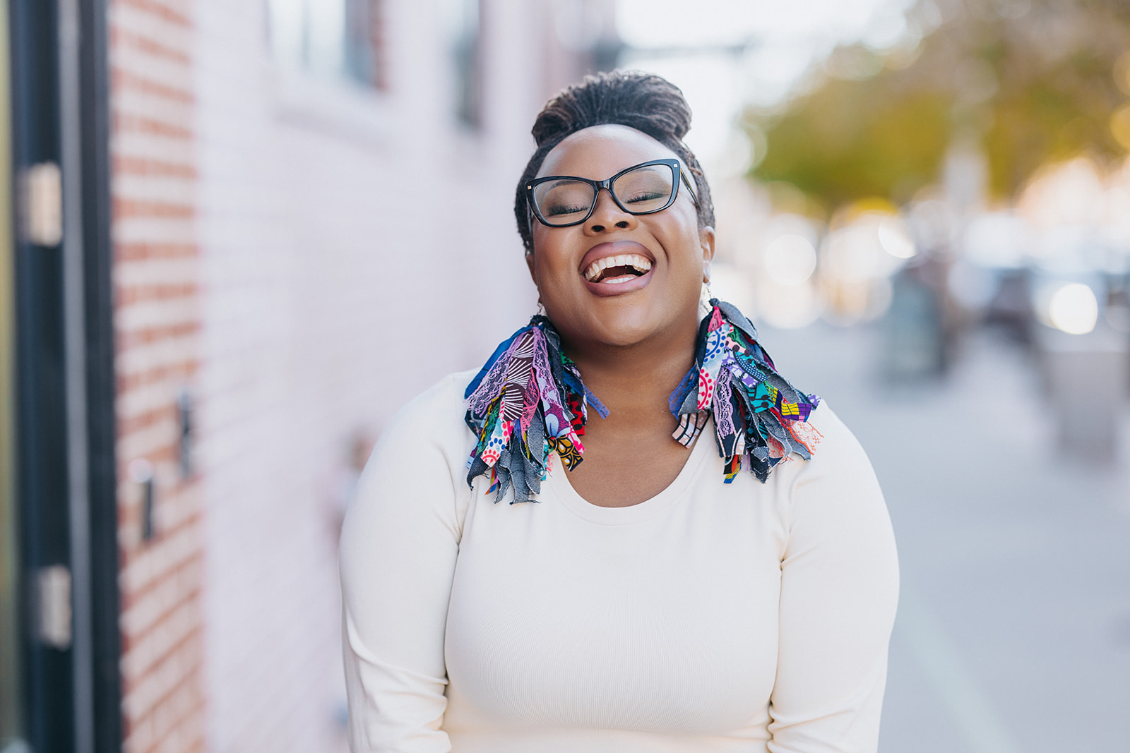 Smiling woman with glasses and colorful dreadlocks outdoors, leaning against a brick wall, sunny day.