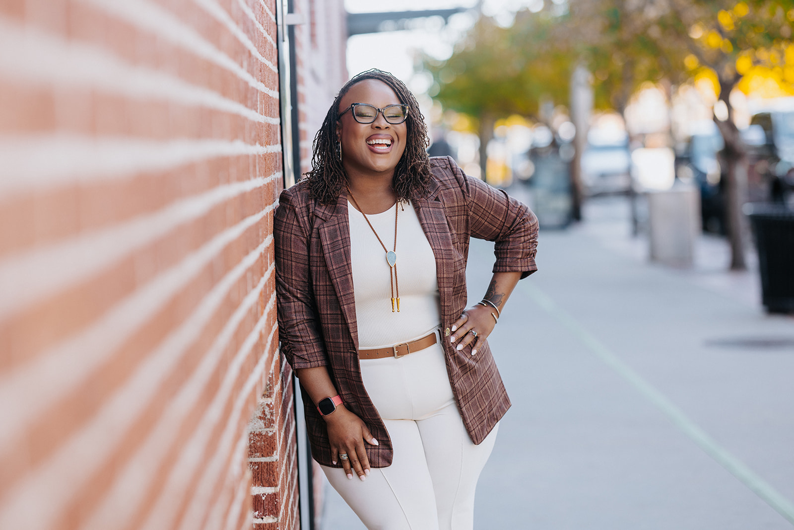 Woman with glasses smiling, standing outdoors next to a brick wall, wearing a brown blazer and white outfit.