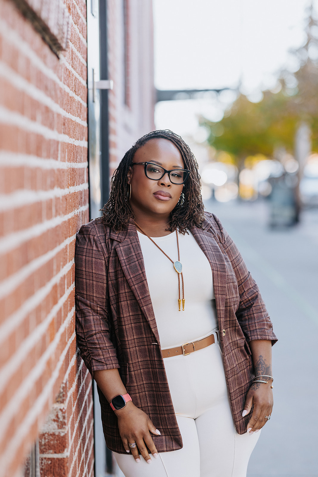 Woman with glasses and curly hair leaning against a brick wall, wearing a plaid blazer and white outfit, outdoors.