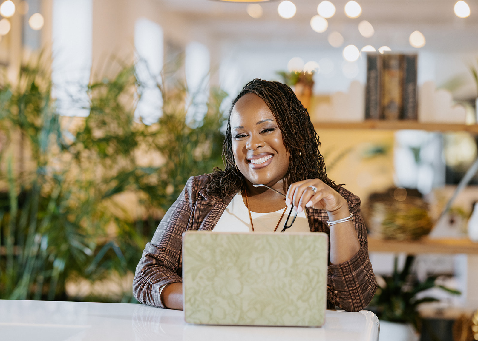 Smiling woman with braided hair using a laptop in a bright, modern space with plants and shelves.