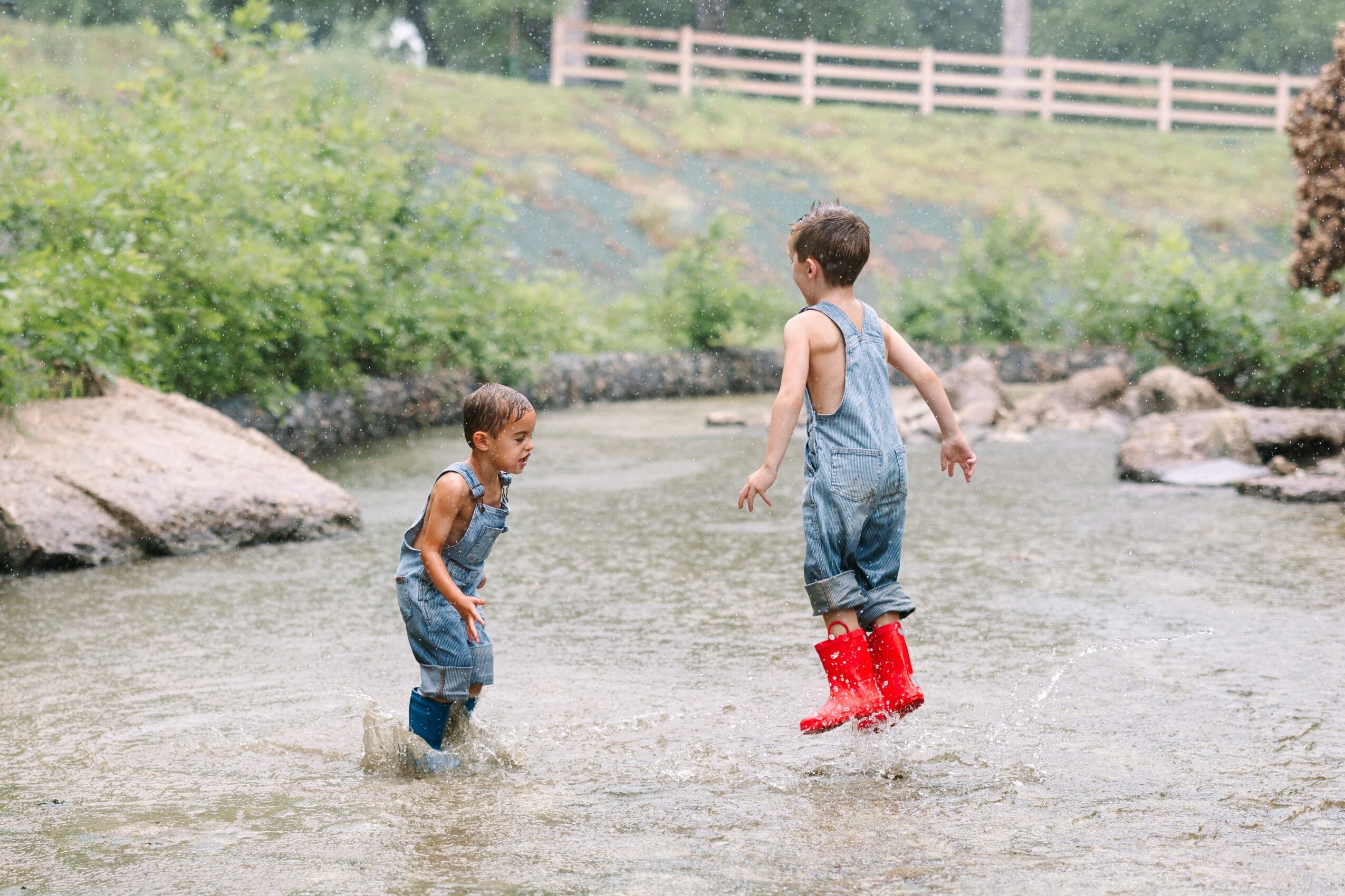 Two children playing in a shallow river, wearing overalls and red boots, with greenery and a fence in the background.