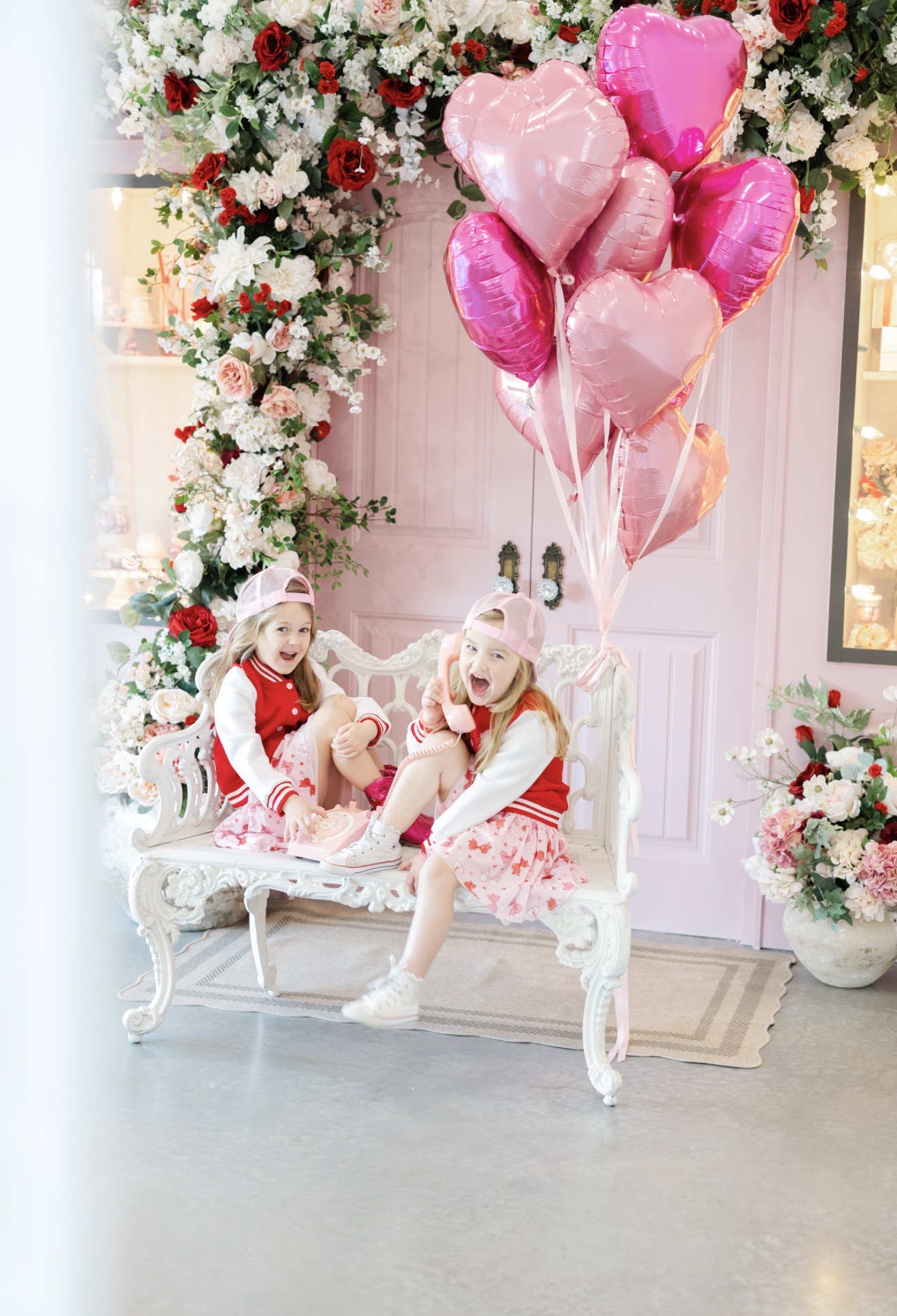 Two children in festive outfits sit on a white ornate bench surrounded by pink and white balloons and floral decorations.