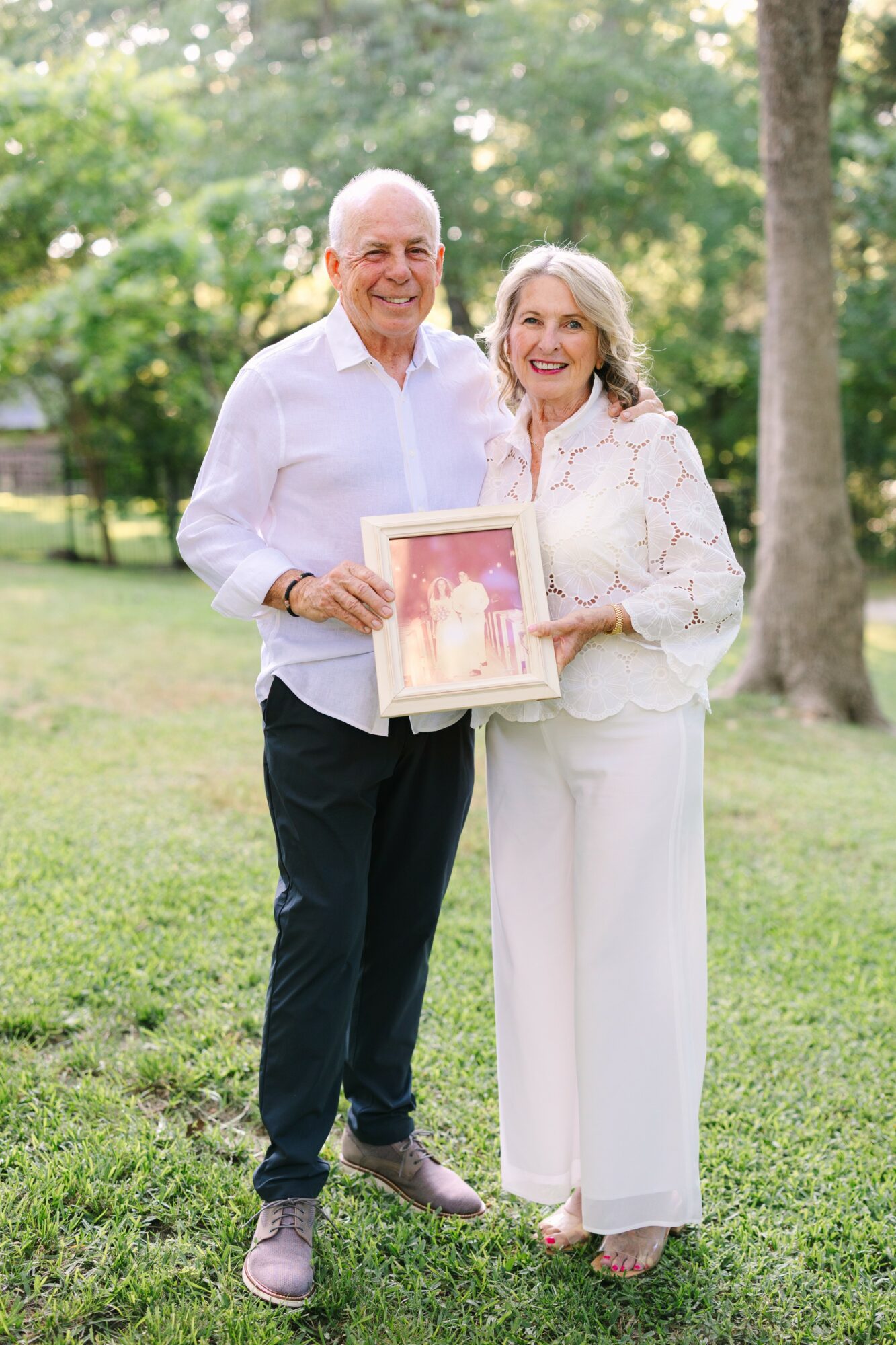 Smiling elderly couple standing outdoors, holding a framed photo, surrounded by trees and greenery.