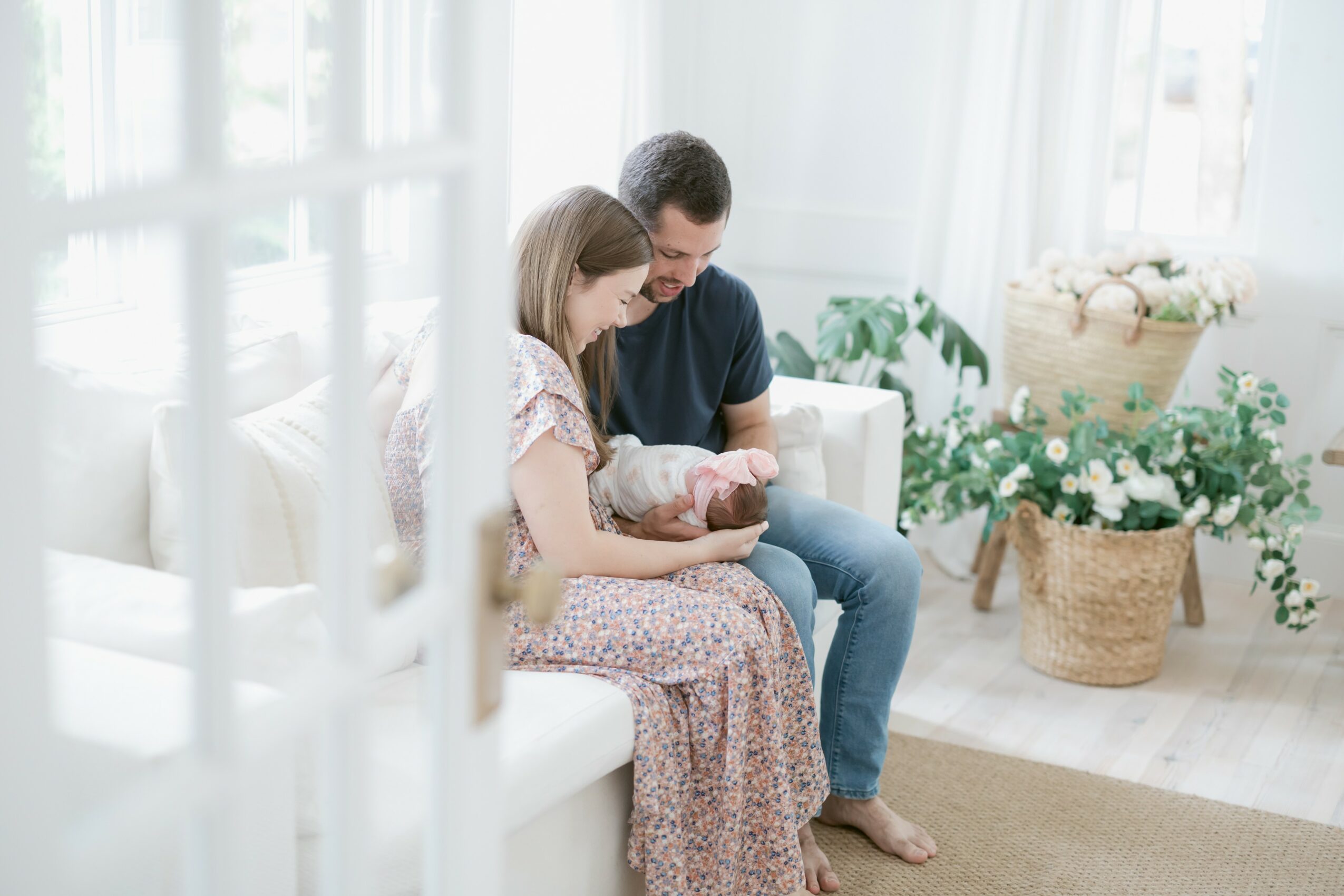 A woman and a man sit on a white sofa holding a newborn baby in a bright, airy room with plants and wicker baskets.