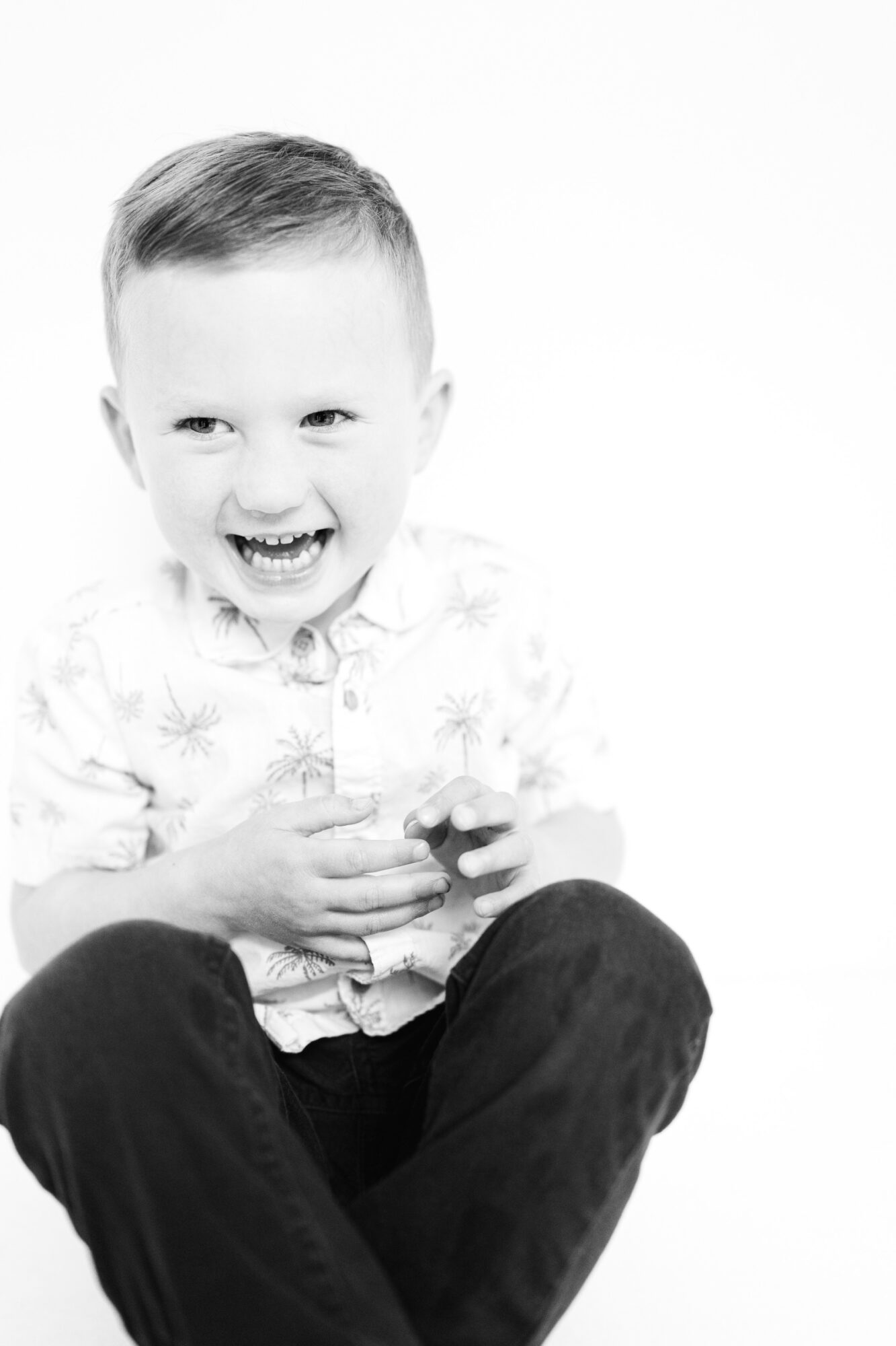Child laughing, sitting with legs crossed, holding an object, wearing a patterned shirt, against a plain background.