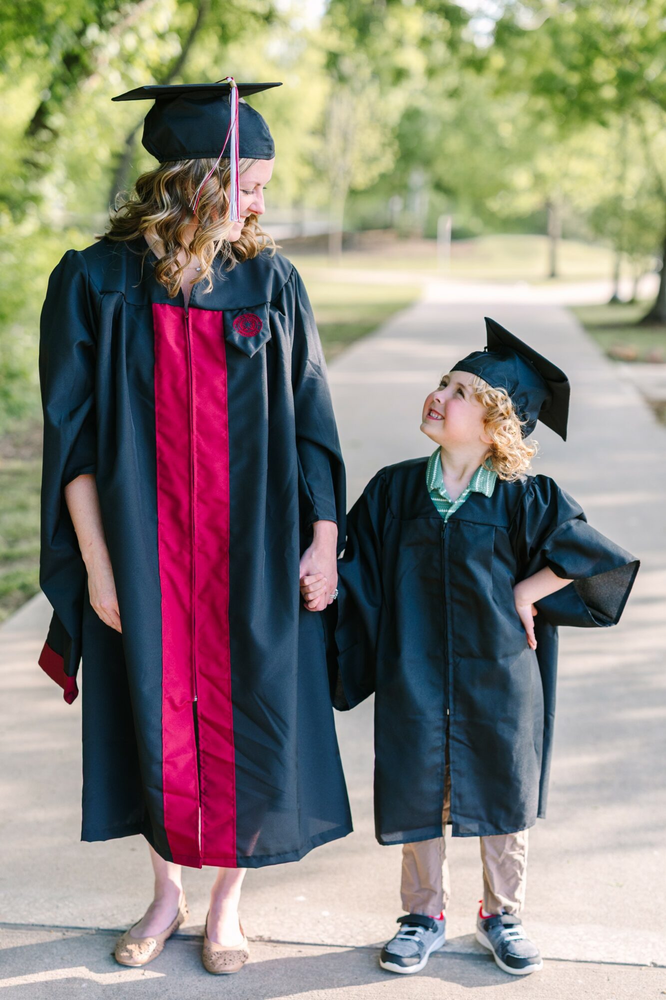 Adult and child in graduation gowns and caps holding hands outdoors, smiling at each other, with trees in background.