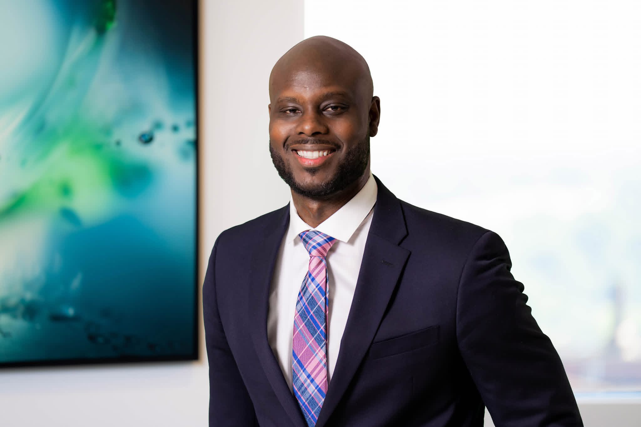 Smiling man in a suit and tie standing indoors with a window and abstract artwork in the background.