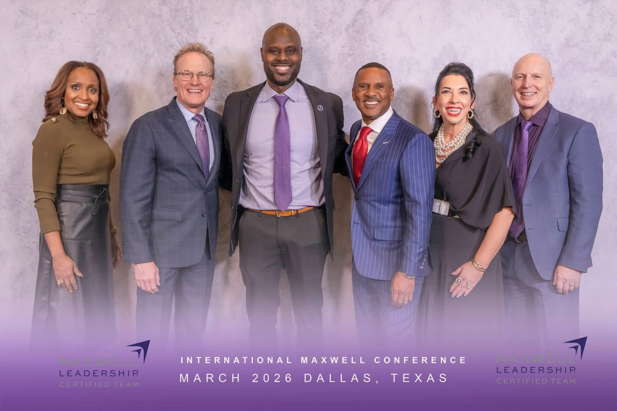 Group of six diverse professionals standing together in front of a gray background, smiling, dressed in business attire.