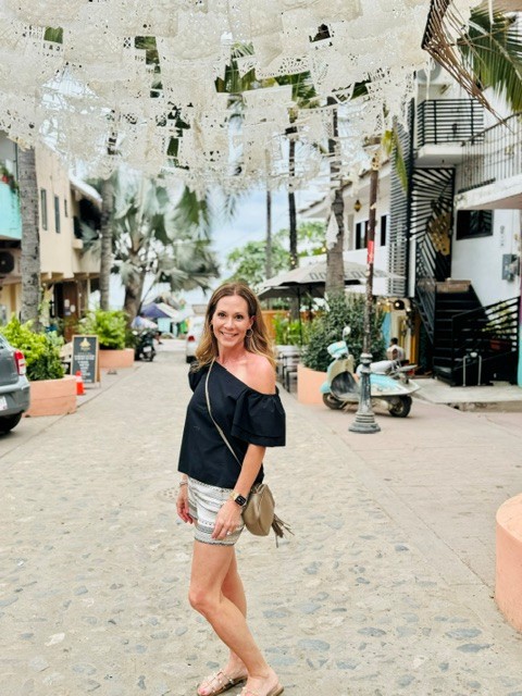 Woman standing on a cobblestone street with hanging decorations overhead, buildings, trees, and parked cars in the background.