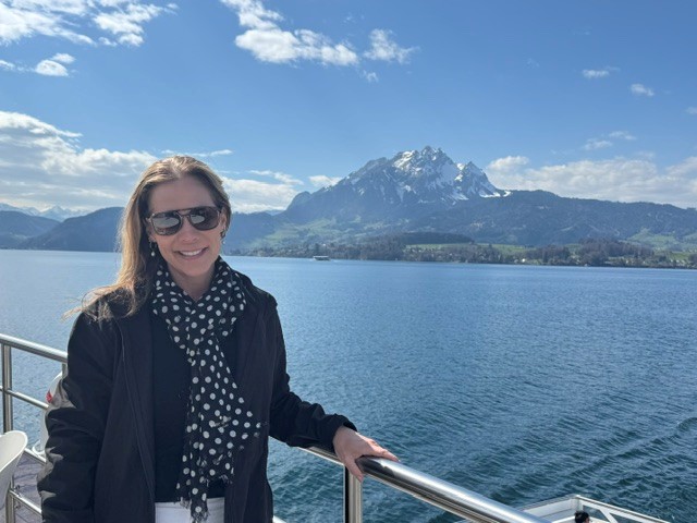 Woman with sunglasses smiling, standing by a railing with a lake and snow-capped mountain in the background.