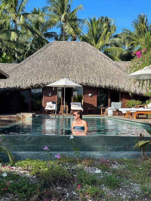 Person swimming in a pool with a thatched roof structure and lush tropical plants nearby.