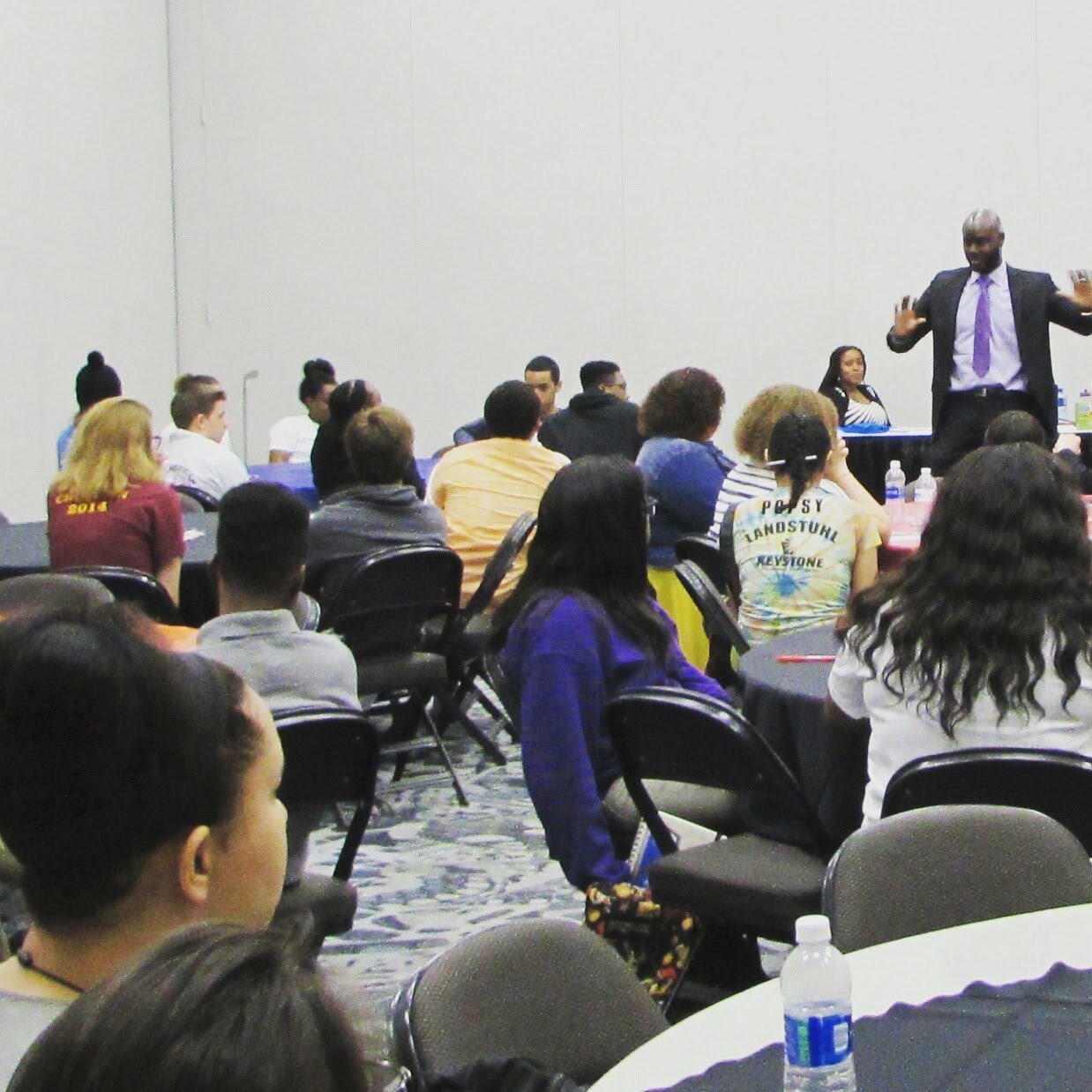 A speaker stands in front of a seated audience in a conference room, gesturing with hands. Audience members face forward, listening attentively.
