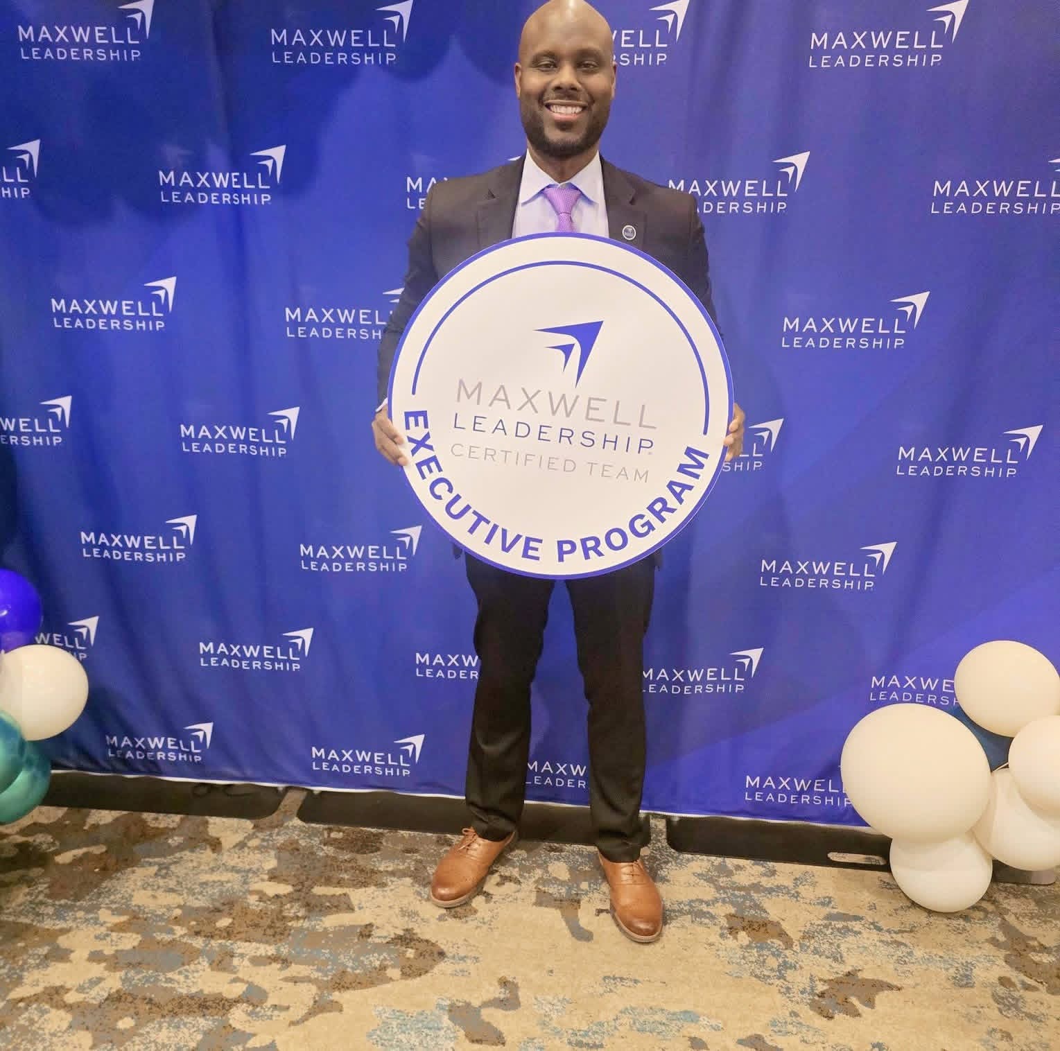 Man in suit holding a large circular sign with Maxwell Leadership Executive Program logo, standing in front of a blue backdrop with similar logos, decorated with white balloons.