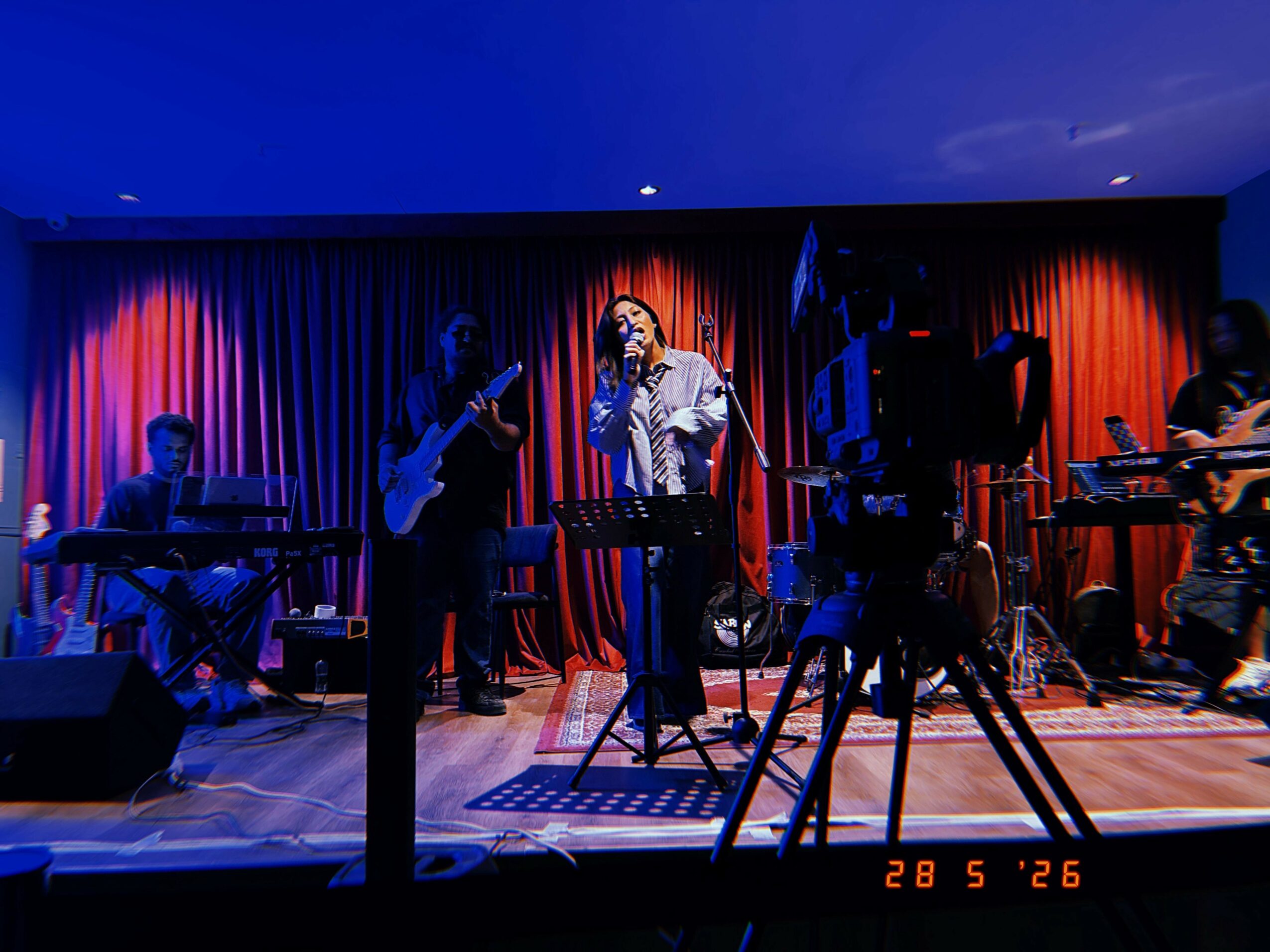 Band performs on stage with red curtains, musician singing into microphone, others playing instruments, blue lighting, wooden floor.