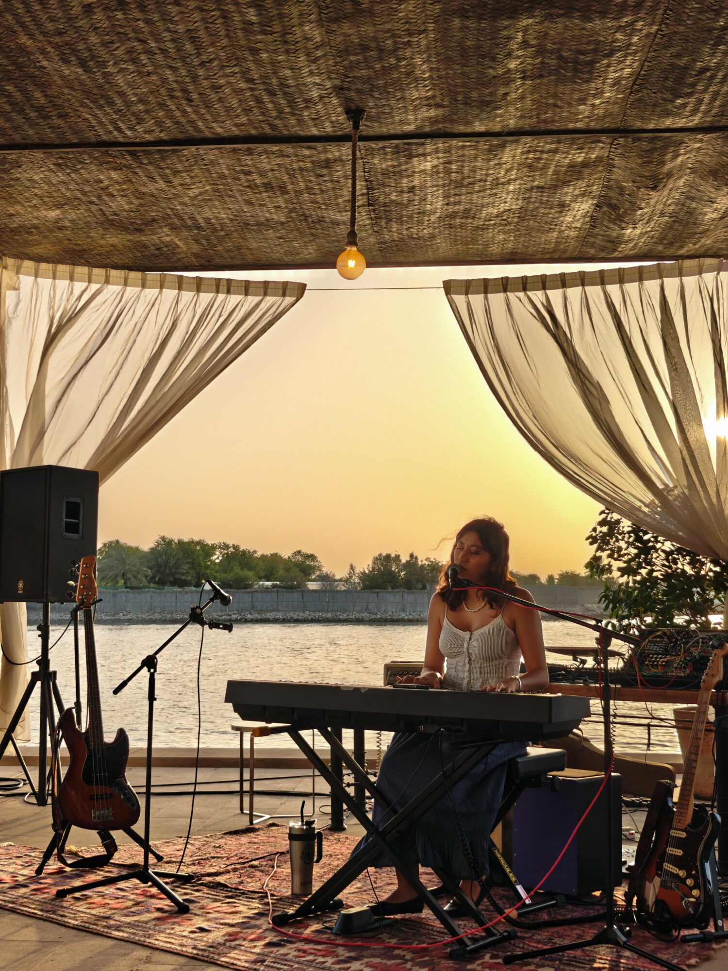 Woman playing keyboard on stage with curtains and water view in background at sunset.
