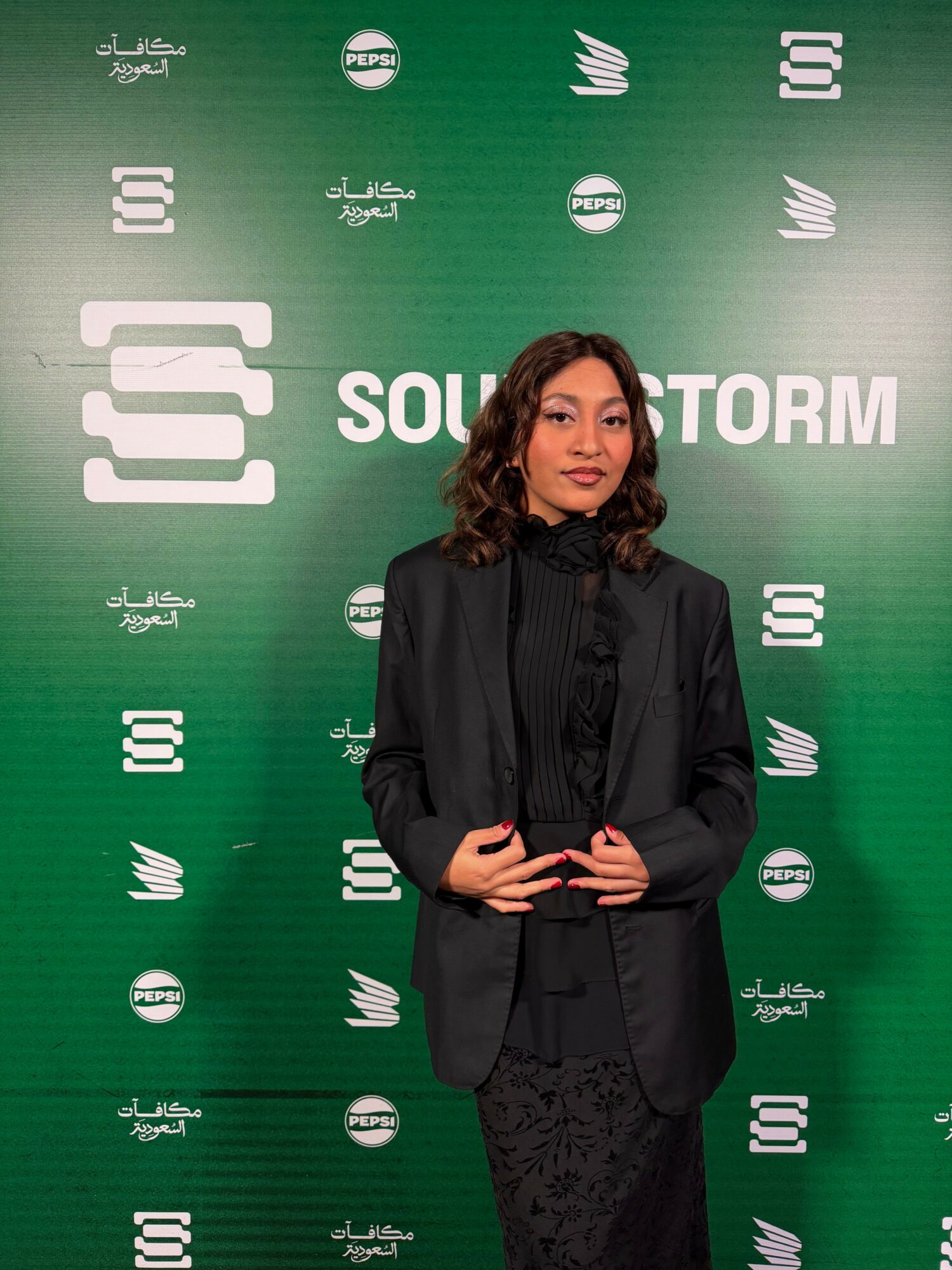 Woman in black outfit standing against green background with logos and text, holding a phone, looking at camera.